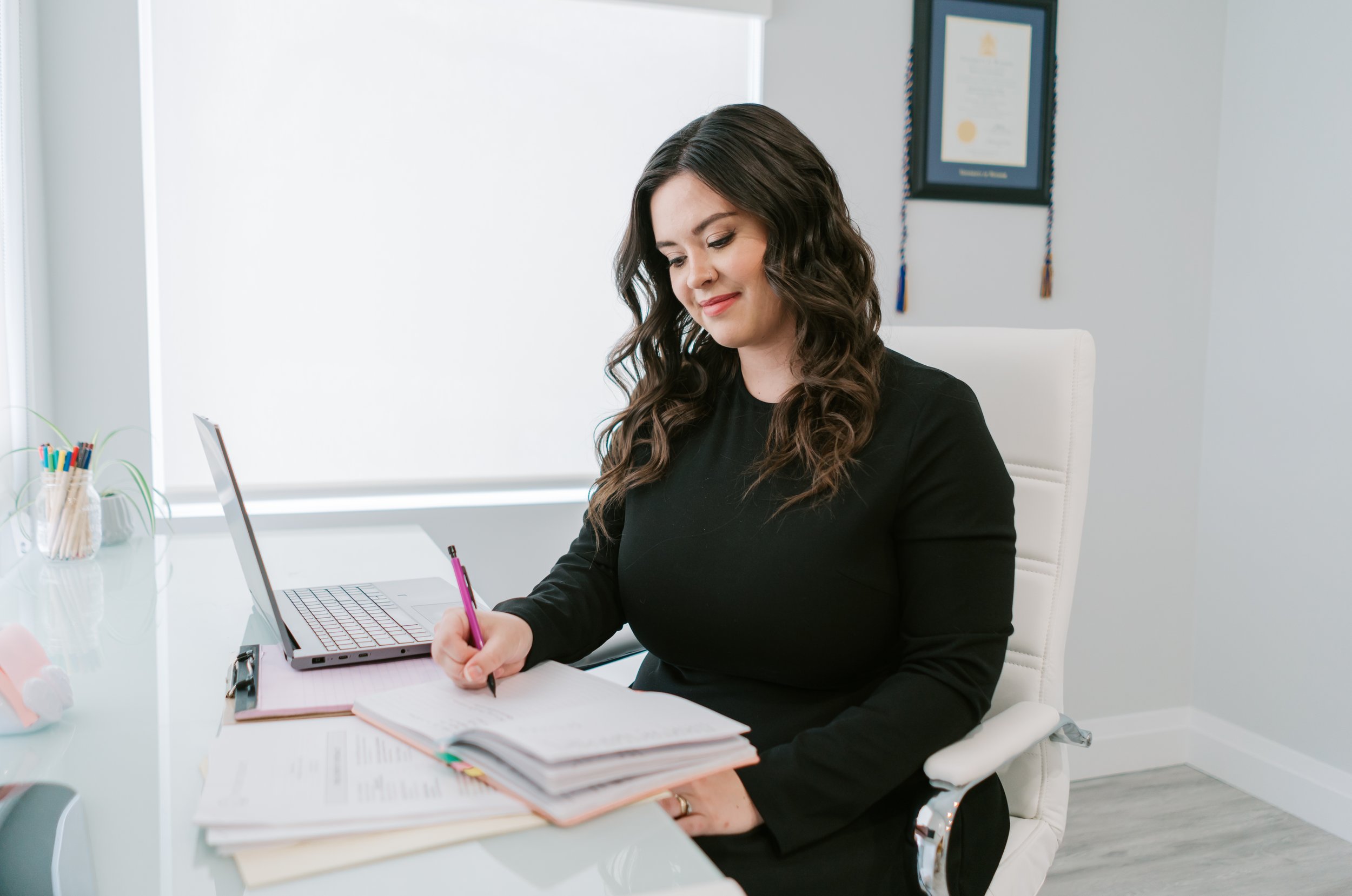 A female therapist with brown curly hair and a blank longsleeved dress sits at a glass desk. A laptop and stack of papers sits in front of her, and she is writing in a notebook.