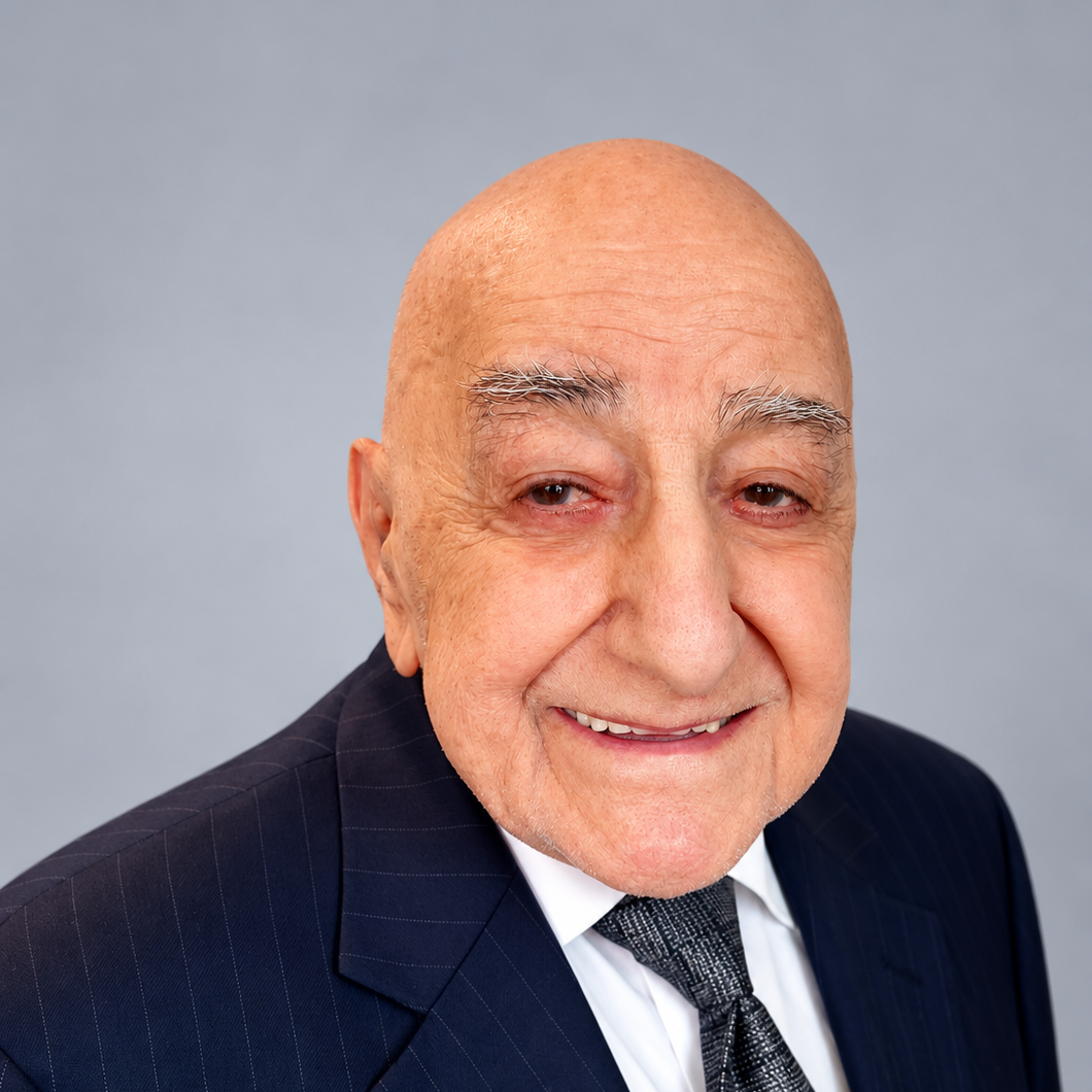 Close-up portrait of an elderly man with a shaved head, gray eyebrows, and a friendly expression, wearing a dark pinstripe suit, white shirt, and patterned tie, against a plain gray background.