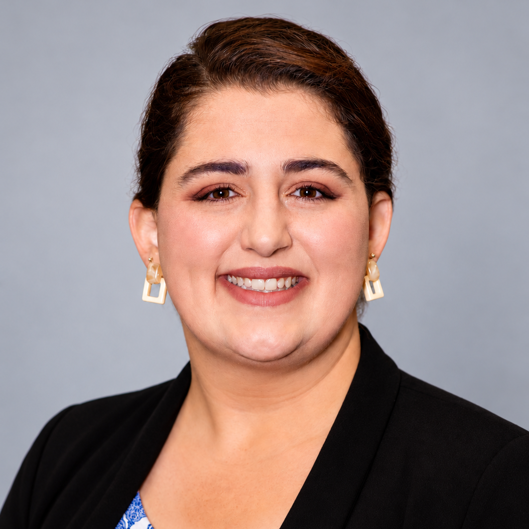A woman with brown hair, wearing earrings and a black blazer, smiling at the camera against a gray background.