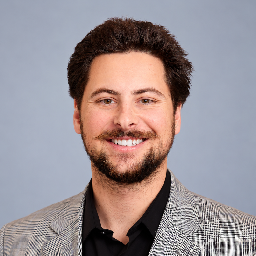 Headshot of smiling man with brown hair, a beard, mustache, wearing a gray checkered suit and black shirt against a plain blue background.