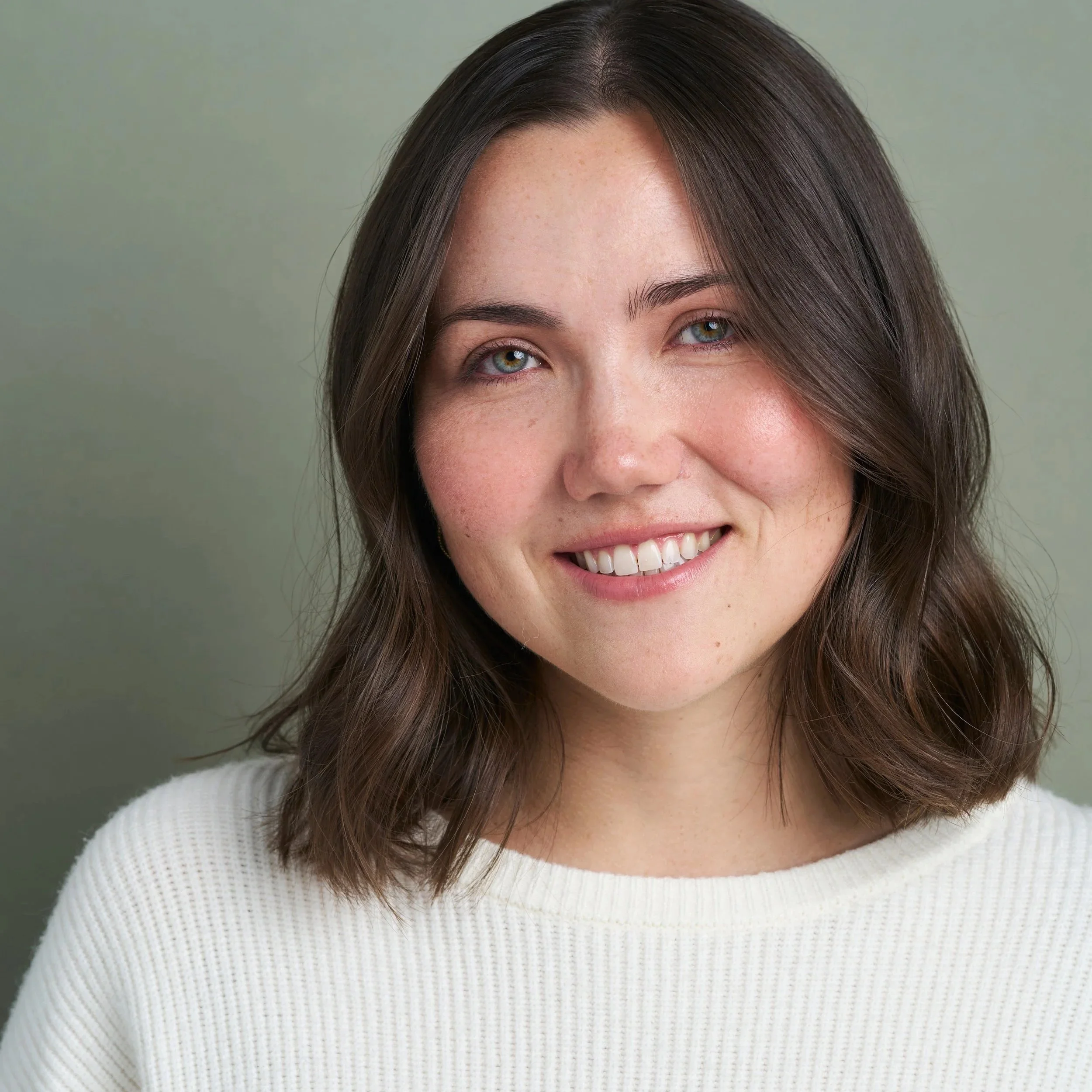Close-up of a smiling woman with brown hair and blue eyes, wearing a white textured sweater, against a plain light green background.