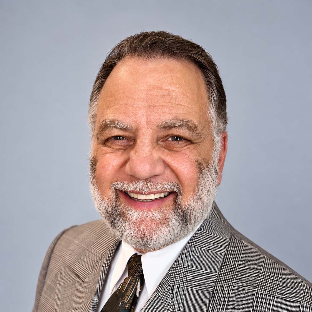 A smiling older man with a beard and gray hair wearing a gray plaid suit, white shirt, and a patterned tie, against a plain light blue background.