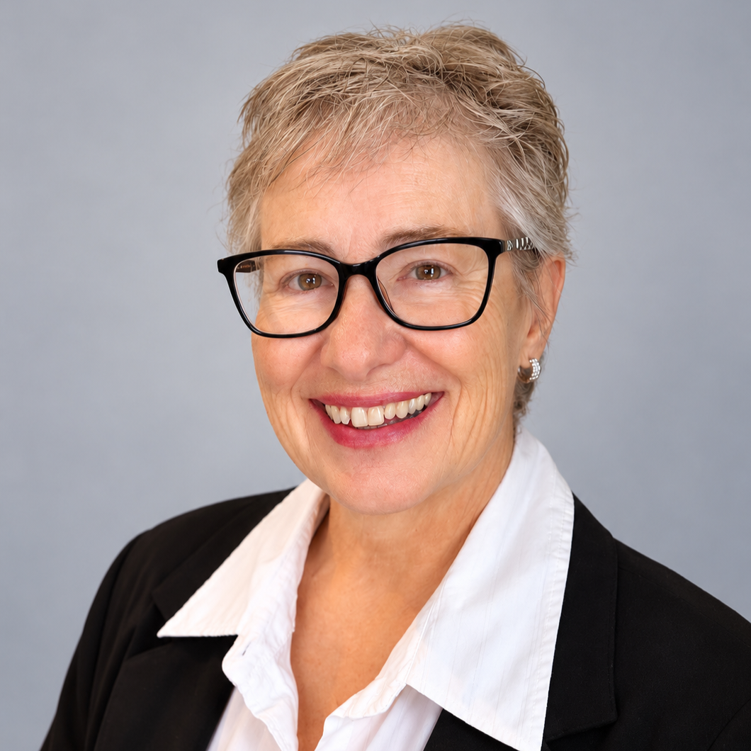 Smiling woman with short gray hair wearing glasses, pearl earrings, a white shirt, and a black blazer against a plain gray background.