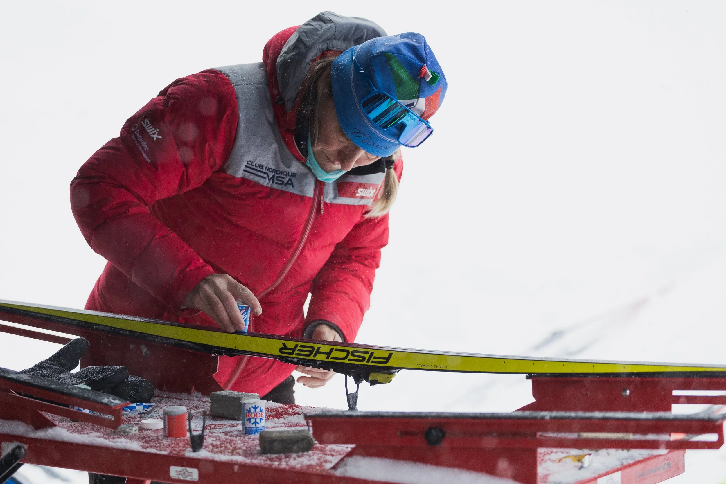 A person dressed in winter gear, including a red jacket and blue goggles, is preparing skis on a ski maintenance station in snowy conditions.