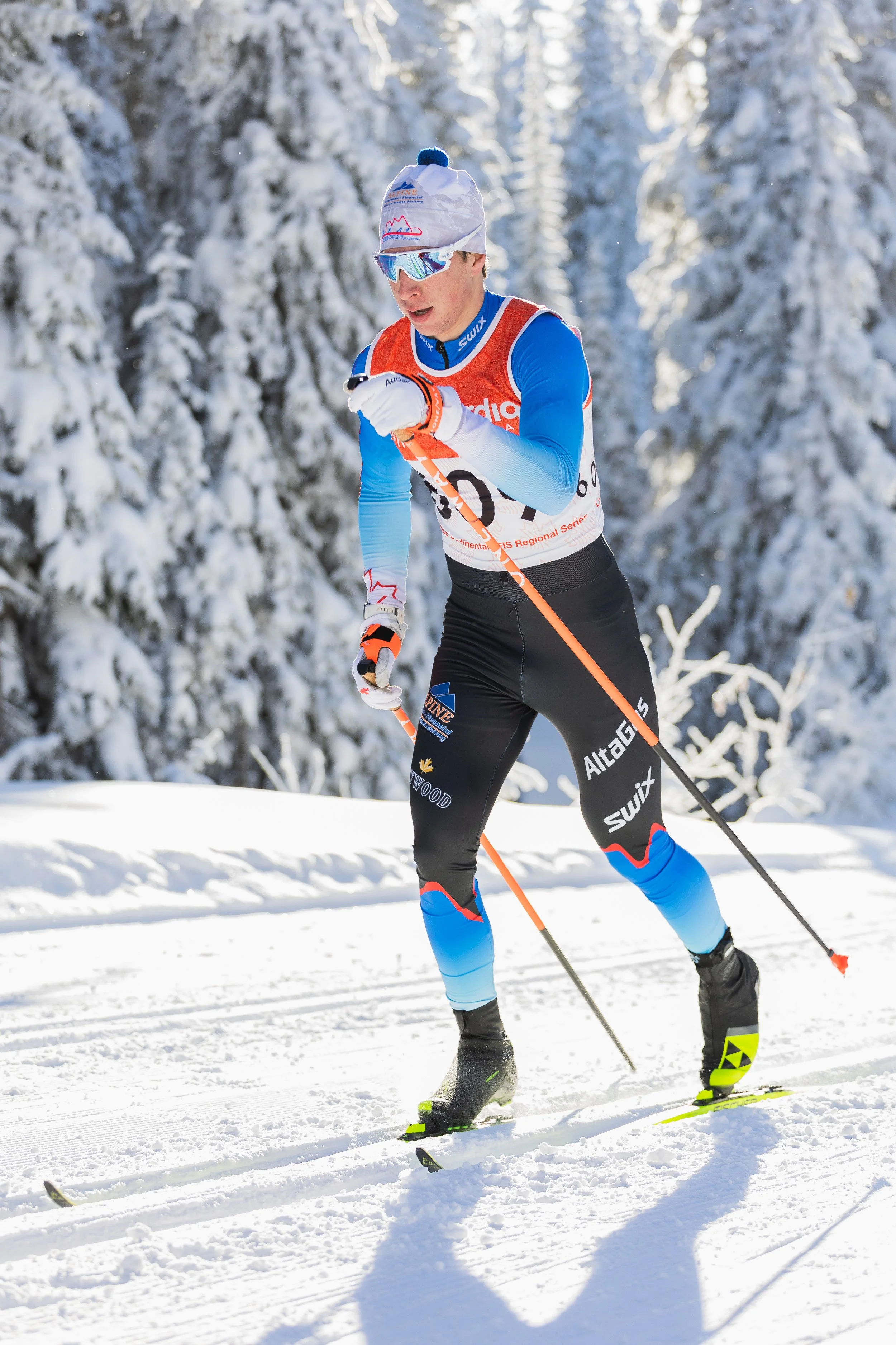 Male cross-country skier in blue and black gear with orange and white bib, wearing sunglasses and a white hat with a pom-pom, skiing on snowy trail surrounded by snow-covered trees.