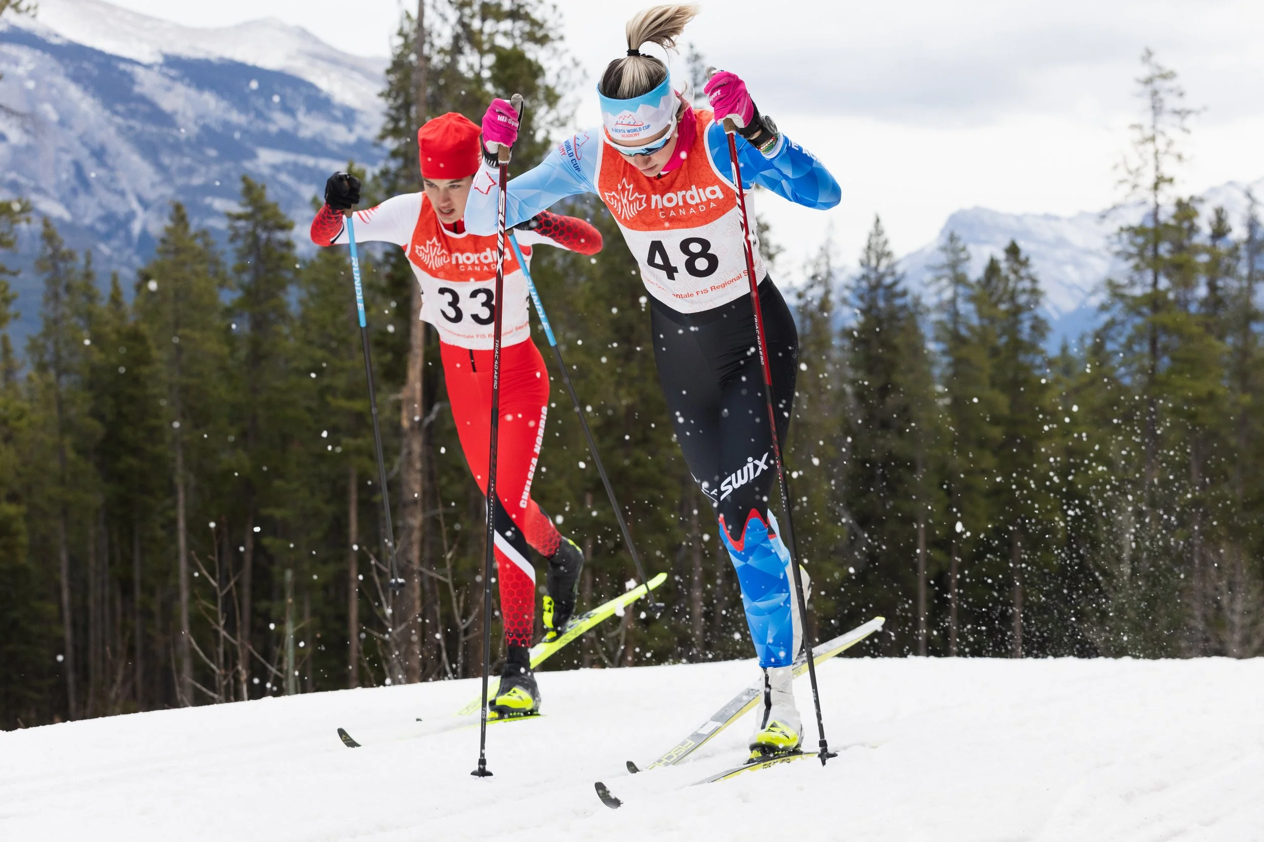 Two female cross-country skiers racing on snowy terrain with a forest and mountains in the background. They are wearing athletic gear, numbered bibs, and using poles as they ski.