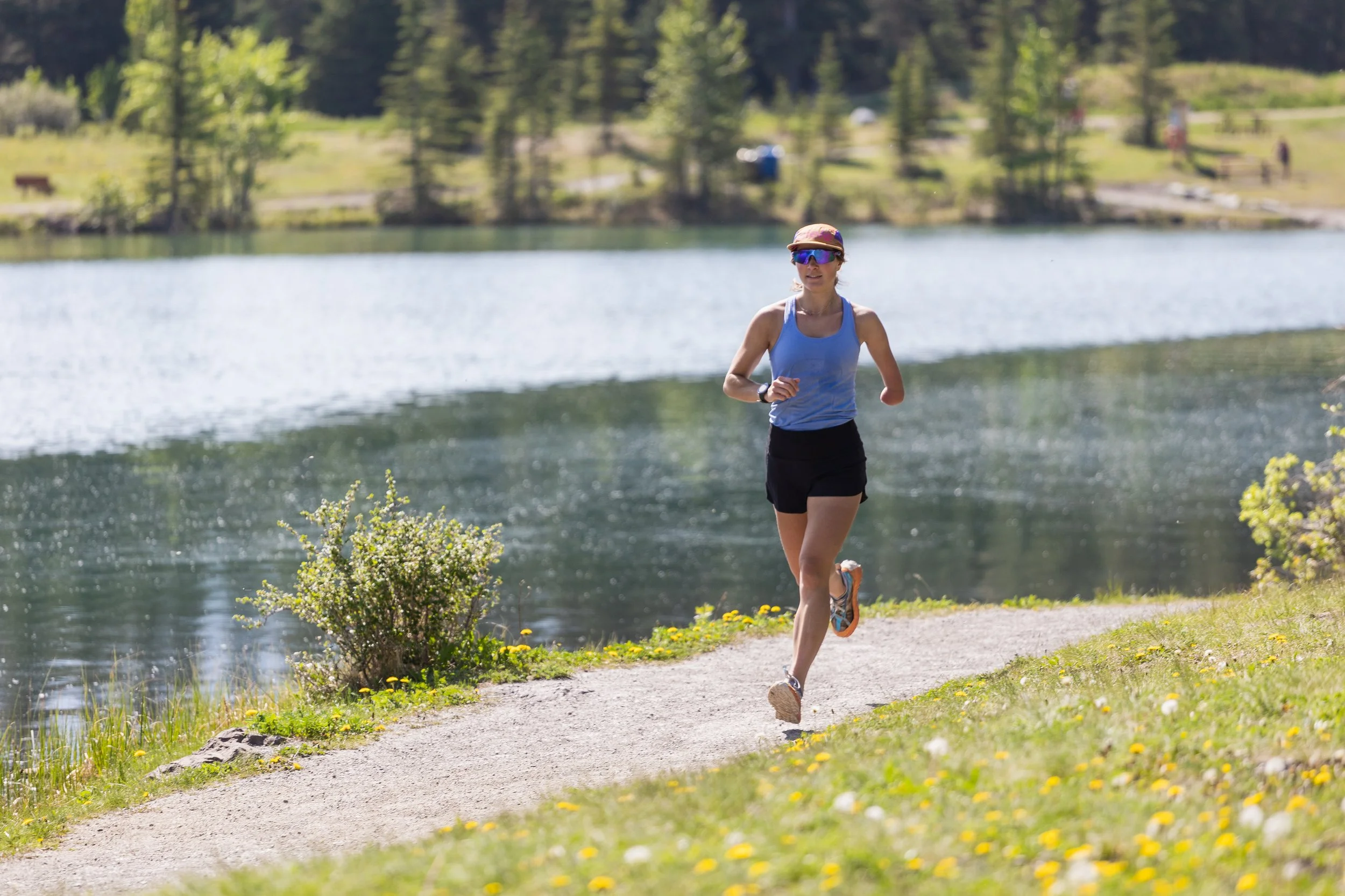 A woman running along a path by a lake, surrounded by trees and greenery on a sunny day.