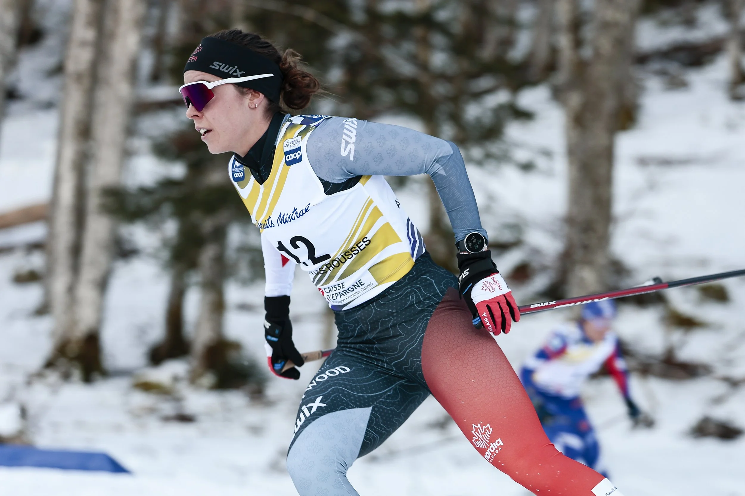 A female cross-country skier in a colorful race suit and sunglasses is skiing through a snowy forested area, with other skiers visible in the background.
