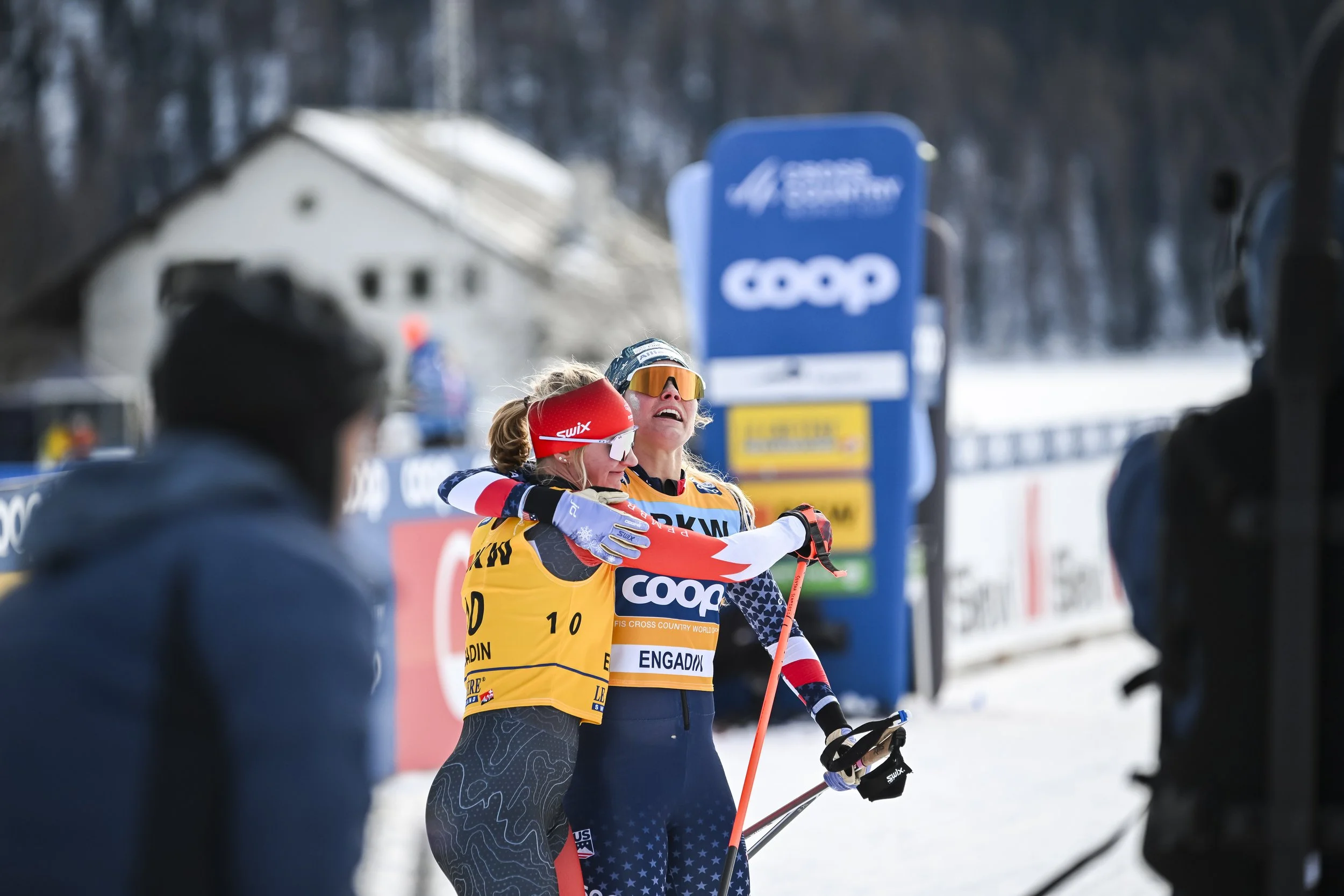 Two female cross-country skiers embracing at a snowy race event, with one wearing a yellow and blue USA team uniform and the other in a patterned black and blue outfit, surrounded by spectators and event banners.
