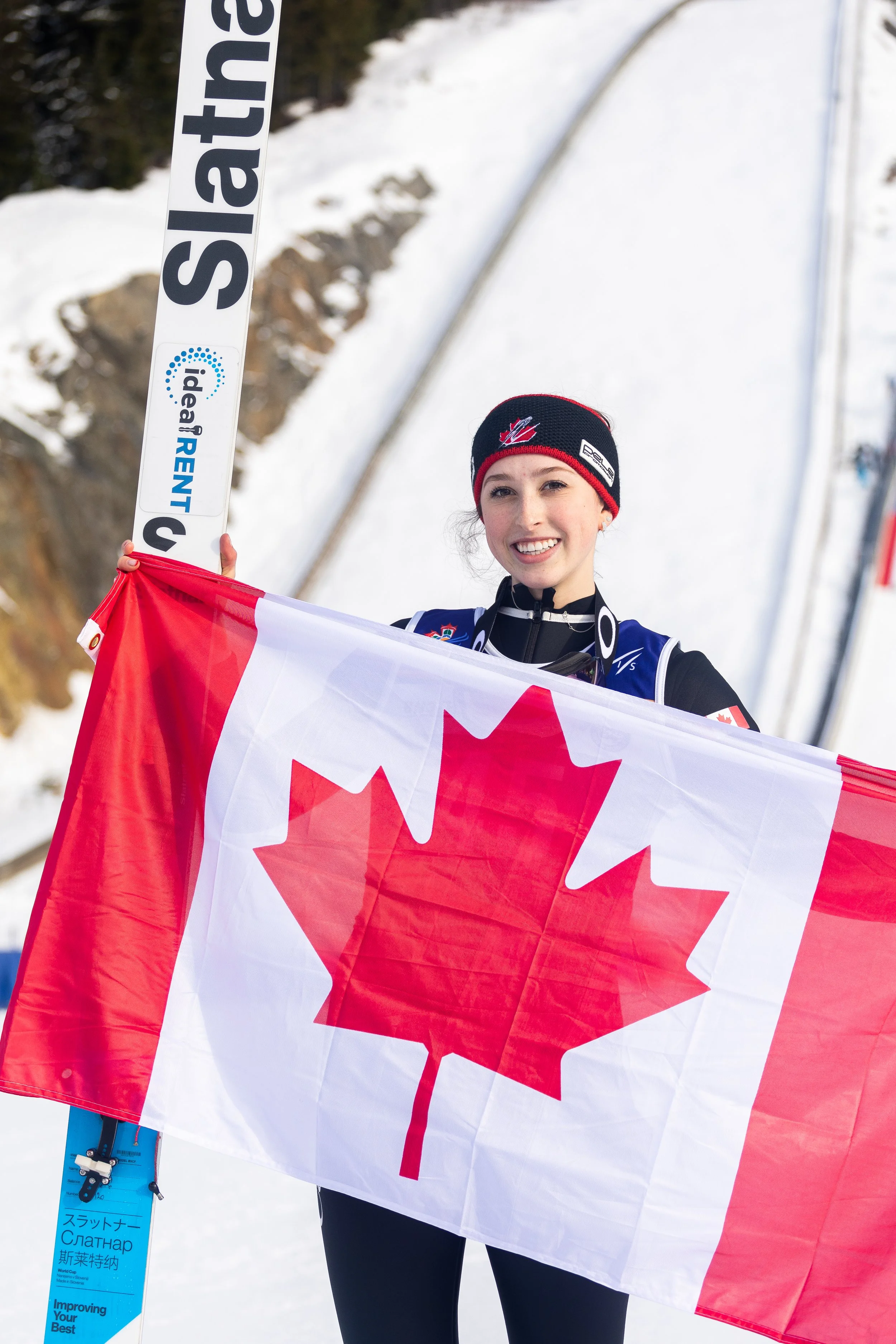 A smiling female skier holding a Canadian flag in a snowy mountain setting.