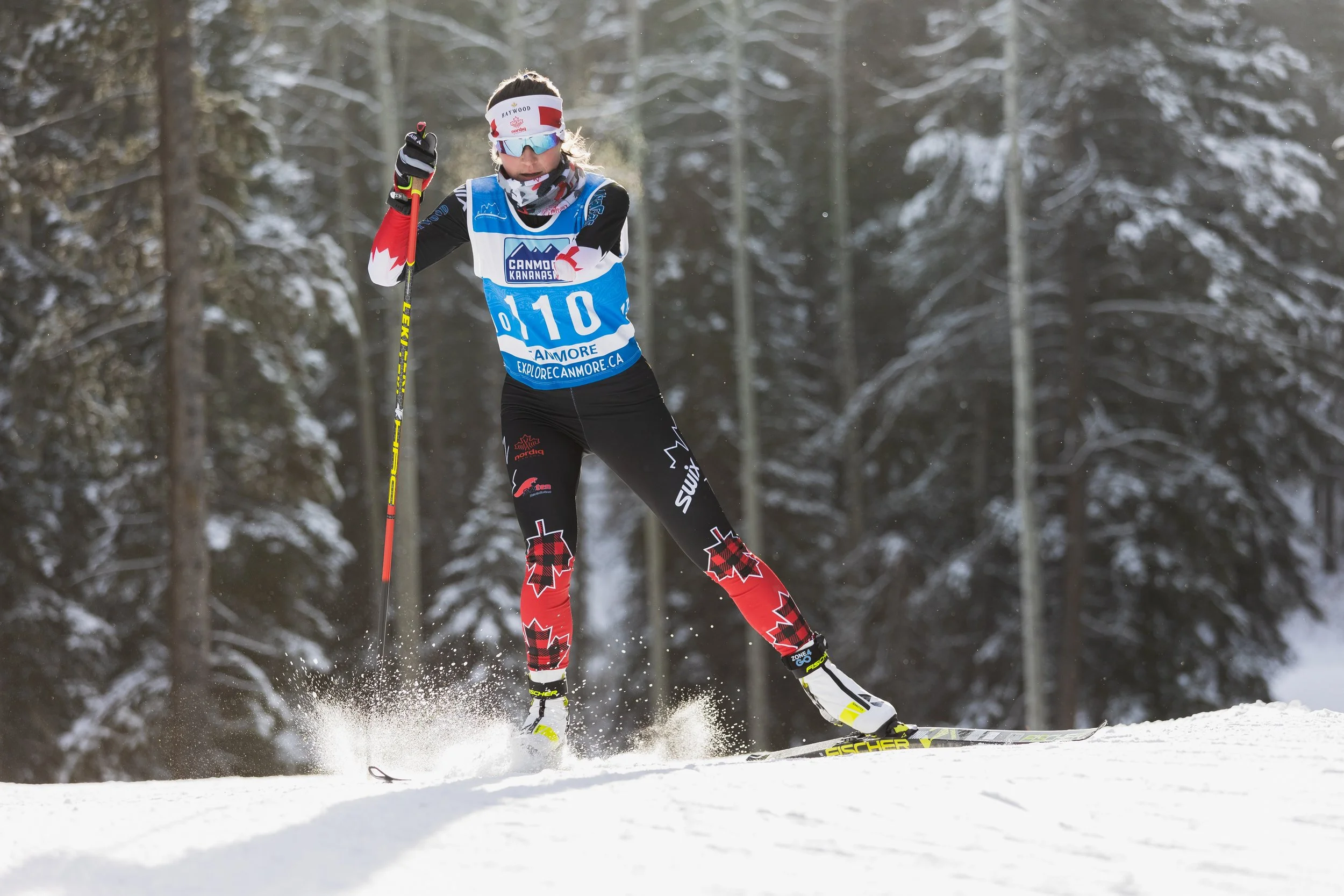 A cross-country skier in winter gear and sunglasses skiing on snow through a forest with snow-covered trees.