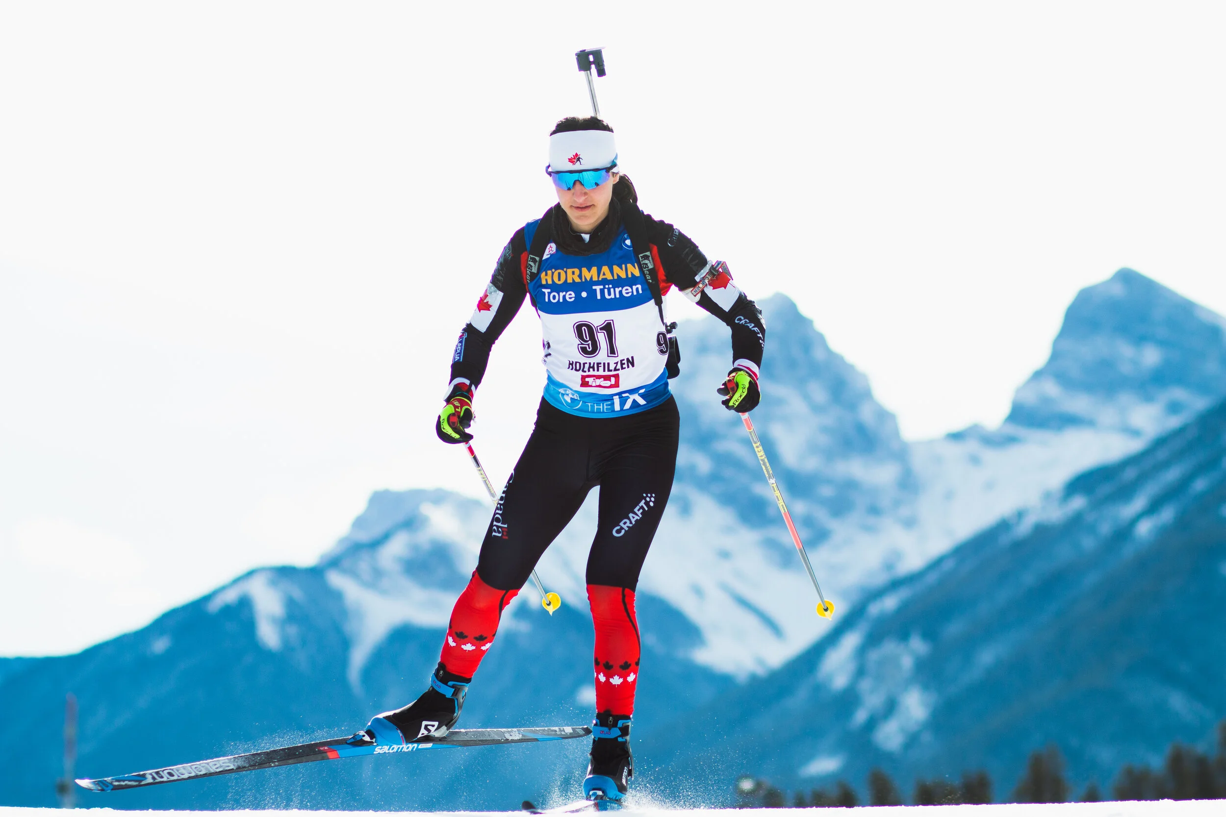 A female cross-country skier wearing sunglasses, a head-mounted camera, a blue and black racing suit with numbered bib, red and black socks, black ski boots, and gloves, skiing on snow with mountain peaks in the background.