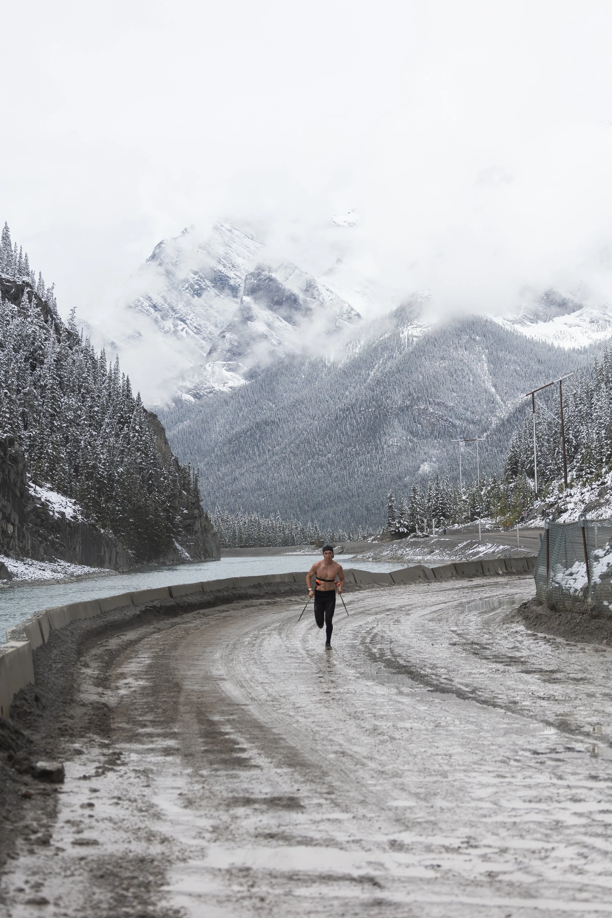 A man is running along a muddy, snow-covered road by a mountain lake surrounded by snow-capped mountains and evergreen trees, with cloudy skies overhead.