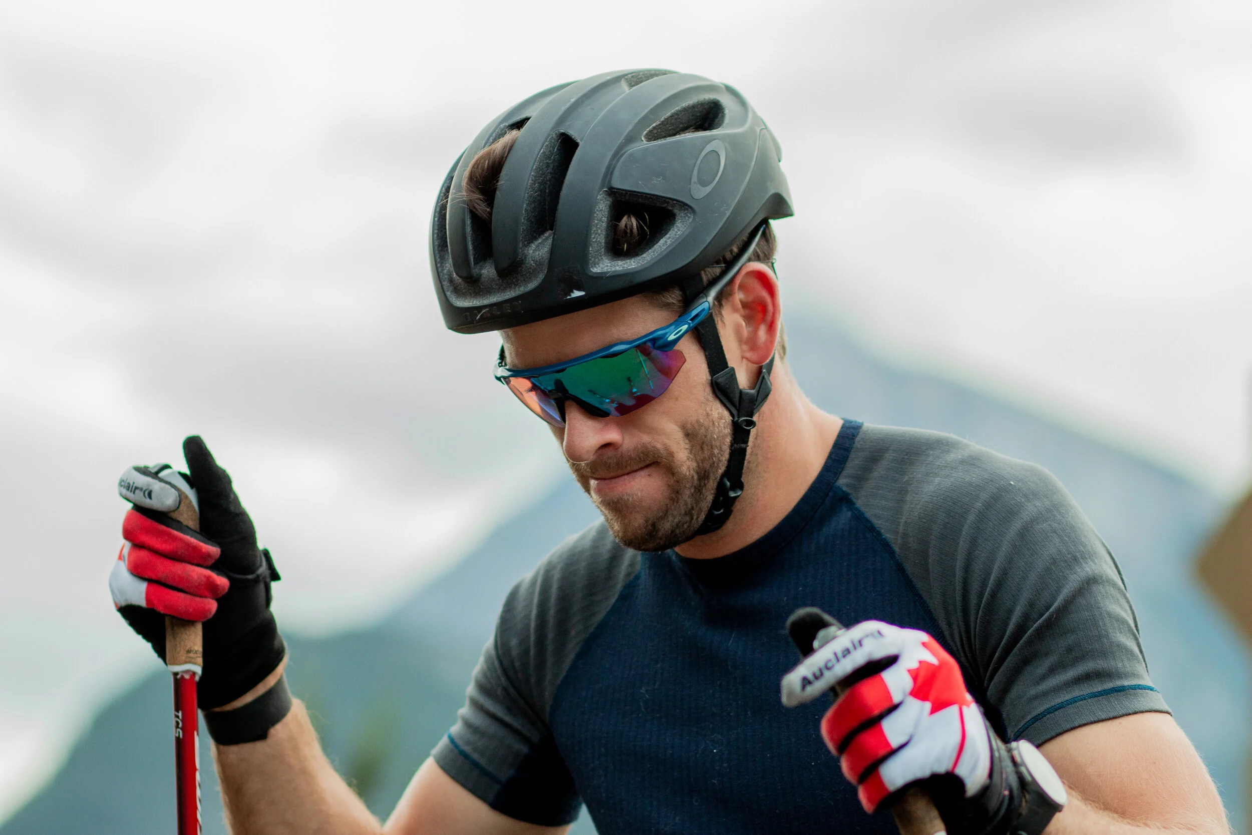 A man wearing a black cycling helmet, reflective sunglasses, and cycling gloves holding a red riding pole, outdoors against a cloudy sky.