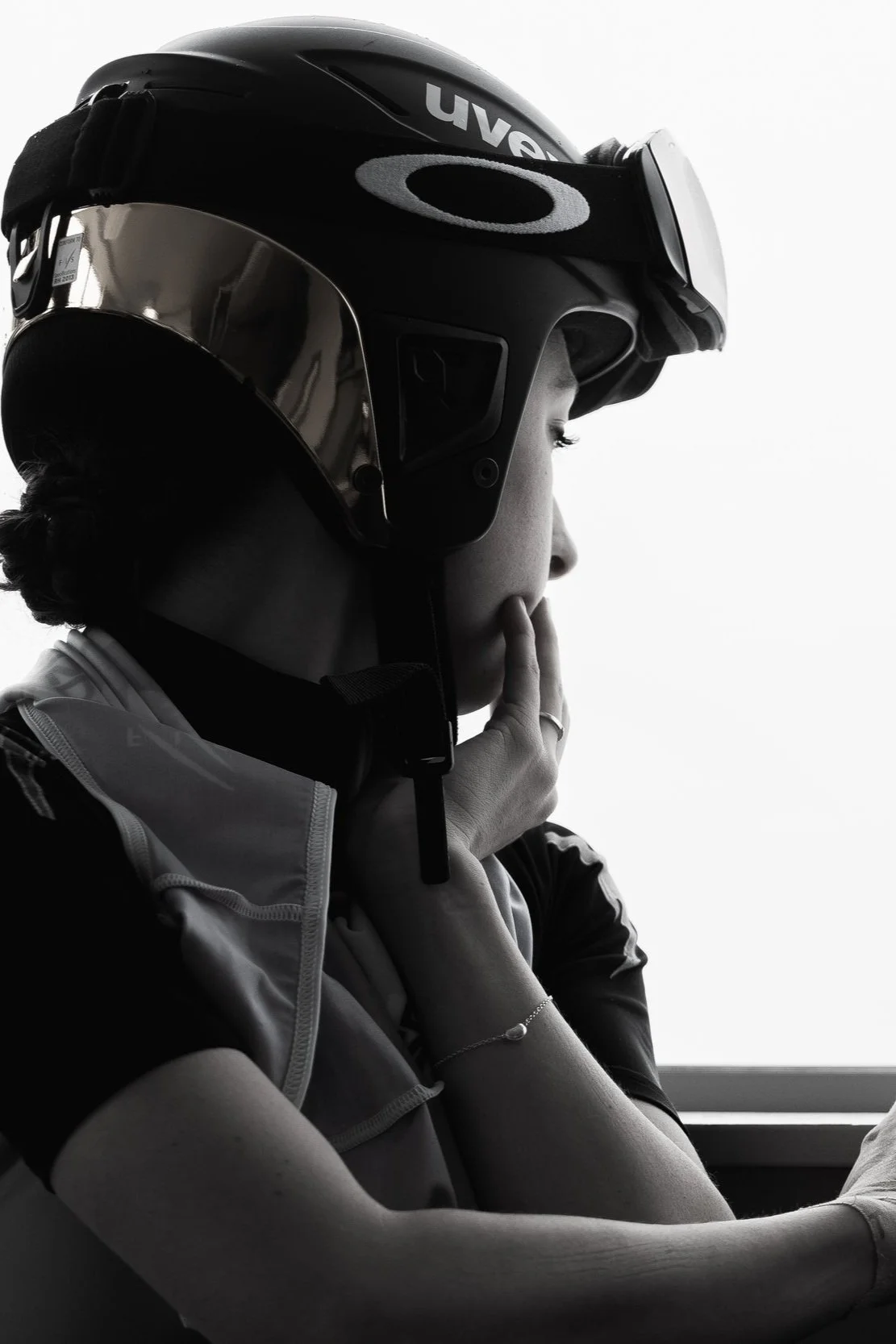 A person wearing a motorcycle helmet and goggles, sitting with their hand near their face, in a black and white photo.