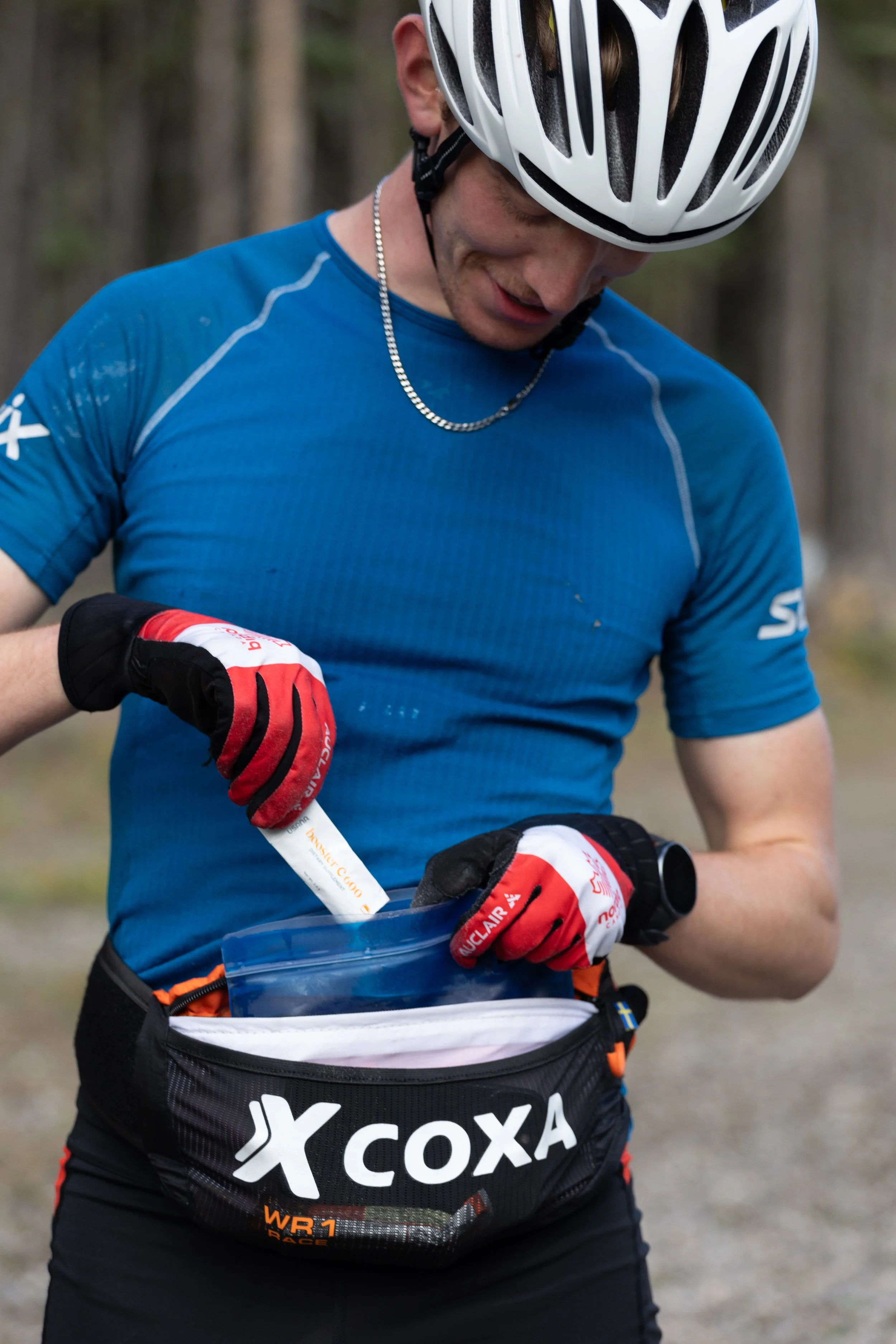A person wearing a cycling helmet and a blue athletic shirt is holding a tube of some substance, possibly energy gel, and standing next to a waist pack with the brand name XCOXA.