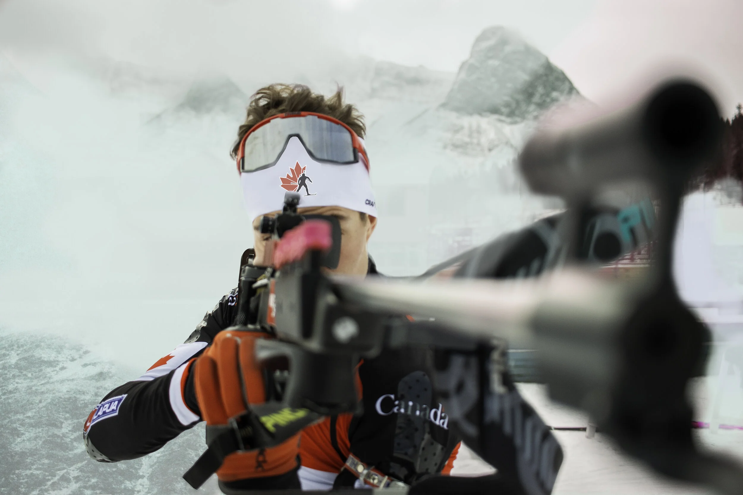 A person in a red and black jacket and Canada-themed headband is snowboarding on a snowy mountain, with mountains in the background and snow spray in the air.