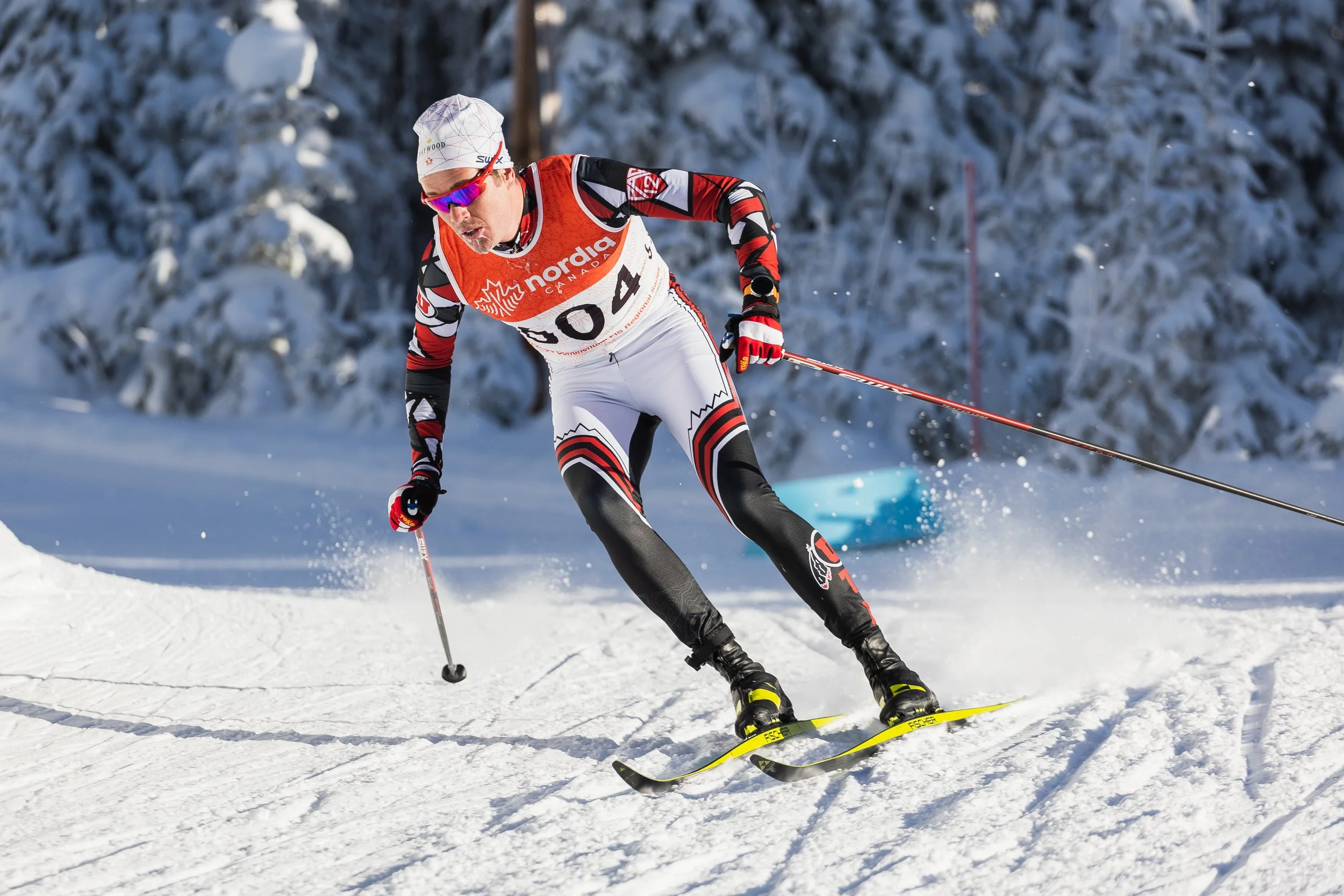 A cross-country skier wearing a white and red racing suit, sunglasses, and a white beanie with colorful designs, skiing through snow with a snowy forest in the background.