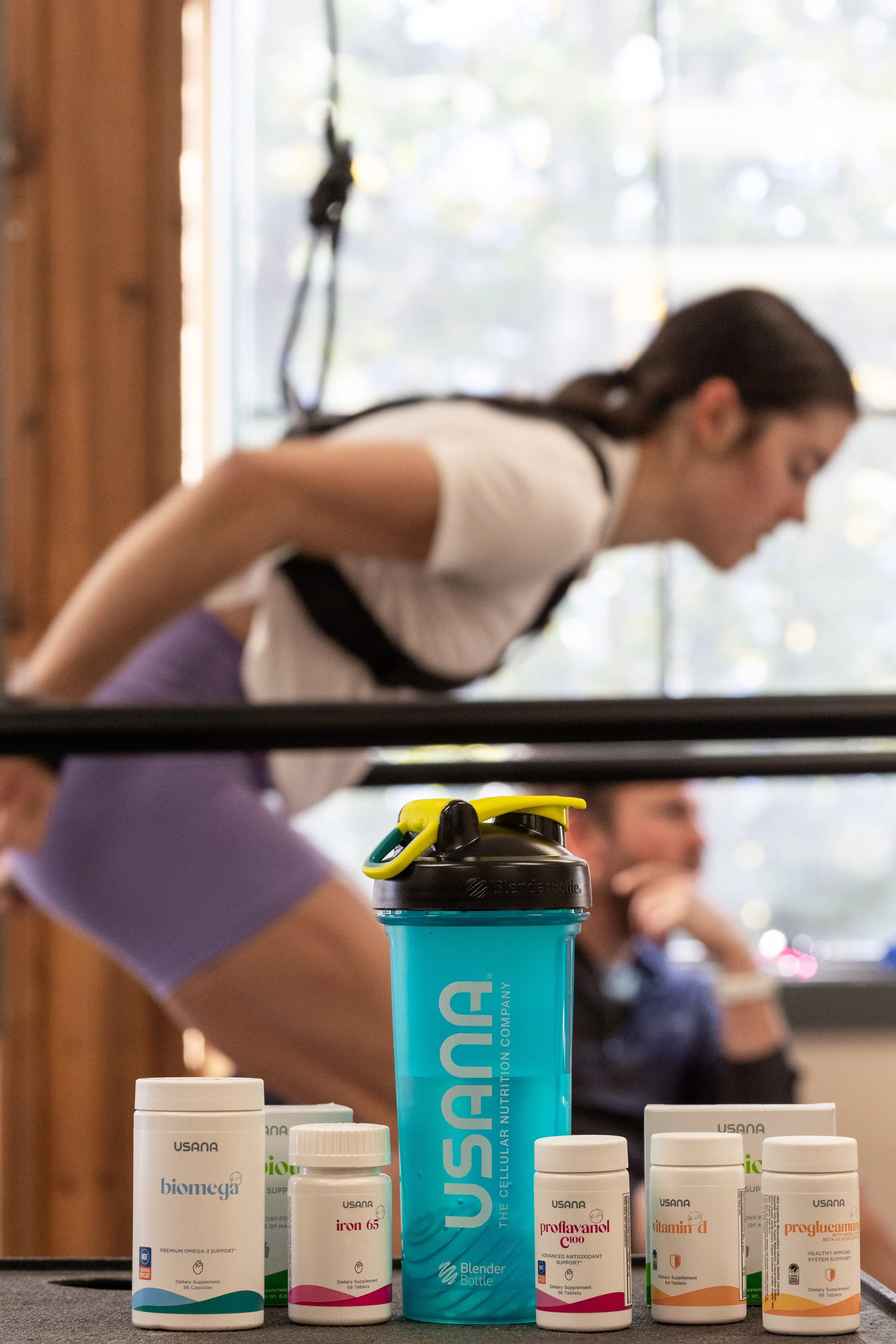 A blue BlenderBottle shaker cup and several bottles of dietary supplements on a table with people in the background.