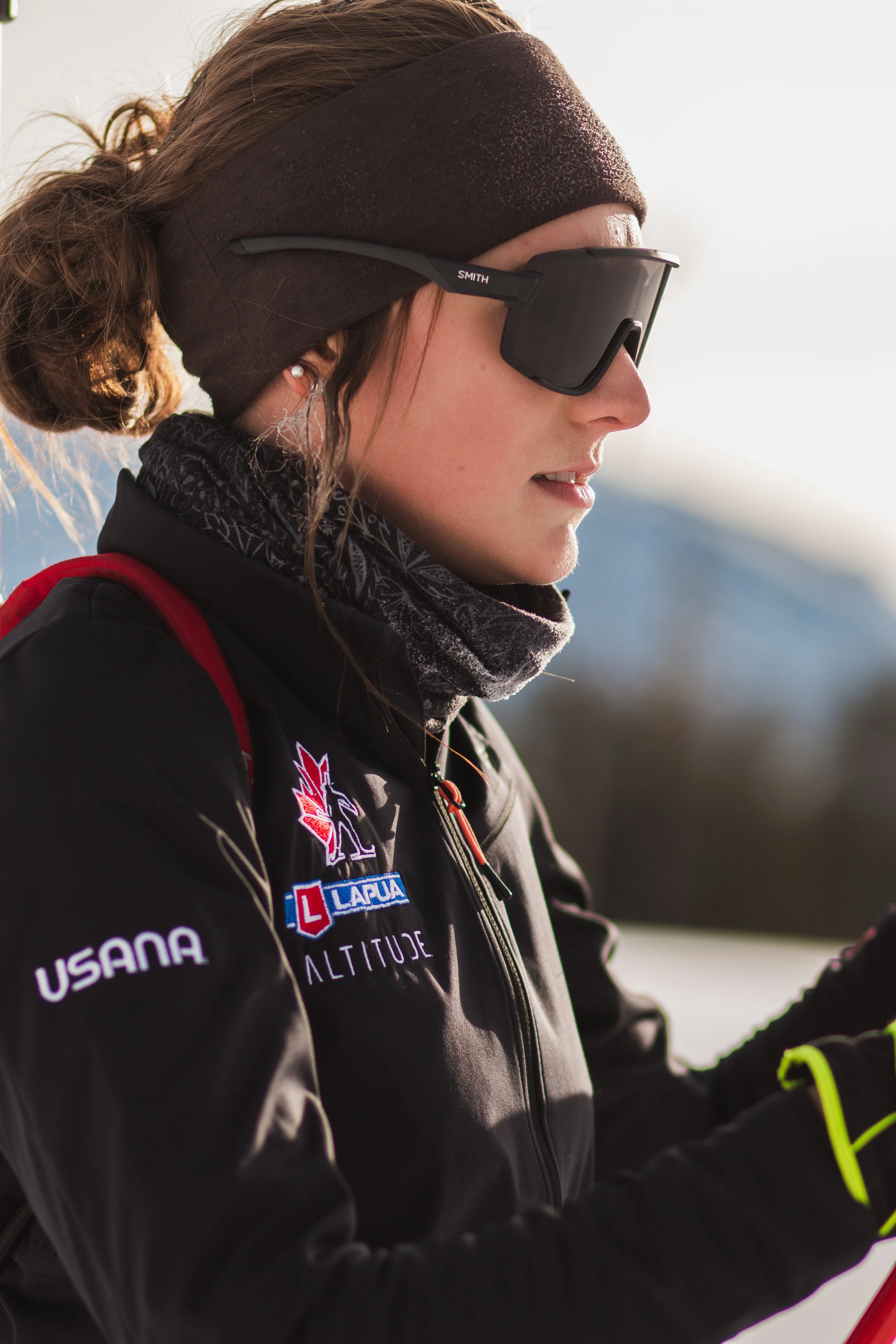 Close-up of a woman wearing a black jacket with several patches, including one saying USA, a black headband, dark sunglasses, and a grey and black neck gaiter, with a snow-capped mountain background.