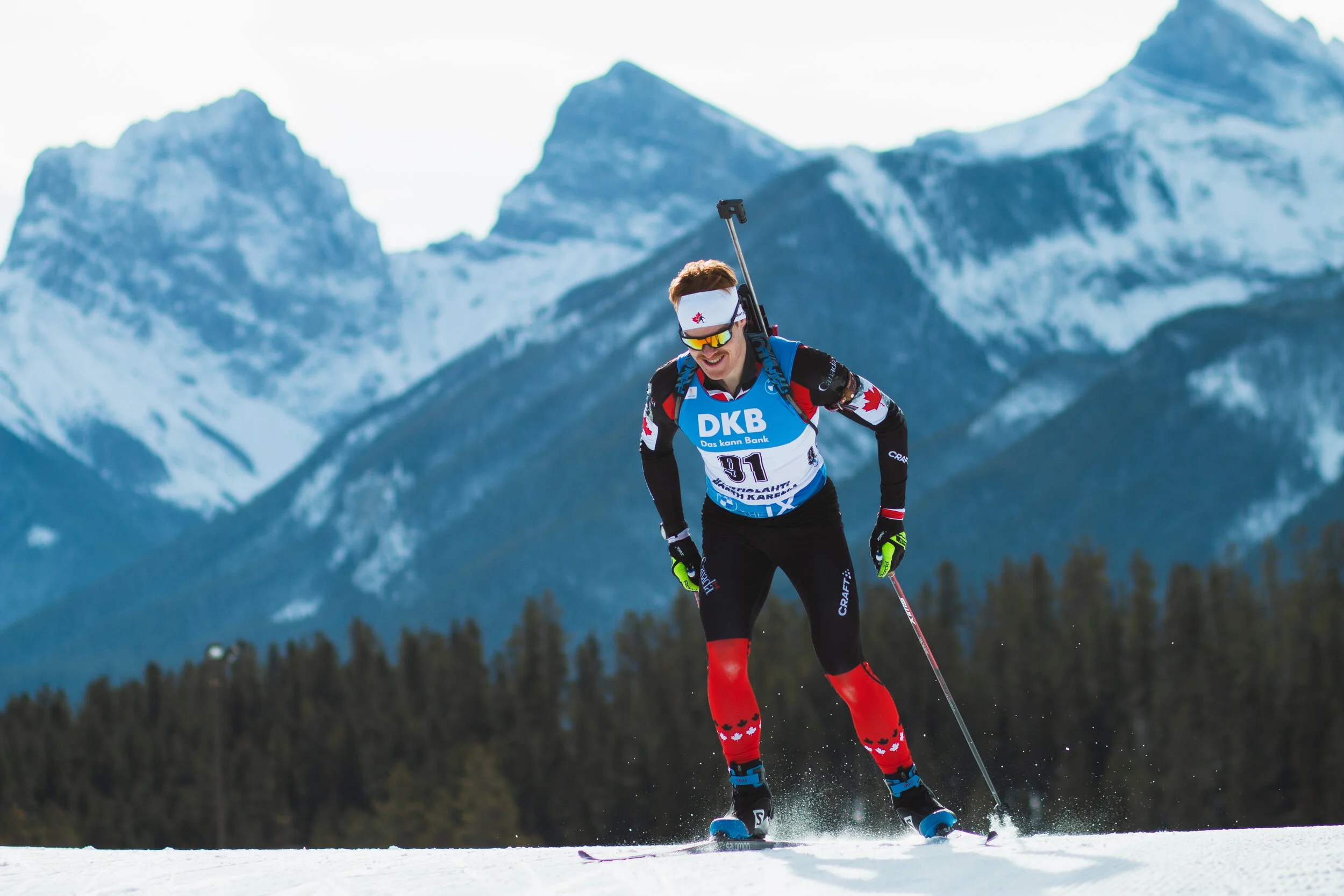 A skier in racing gear, wearing a white headband with a snowflake logo, and racing bib number 91, skiing downhill on snow with mountain peaks in the background.