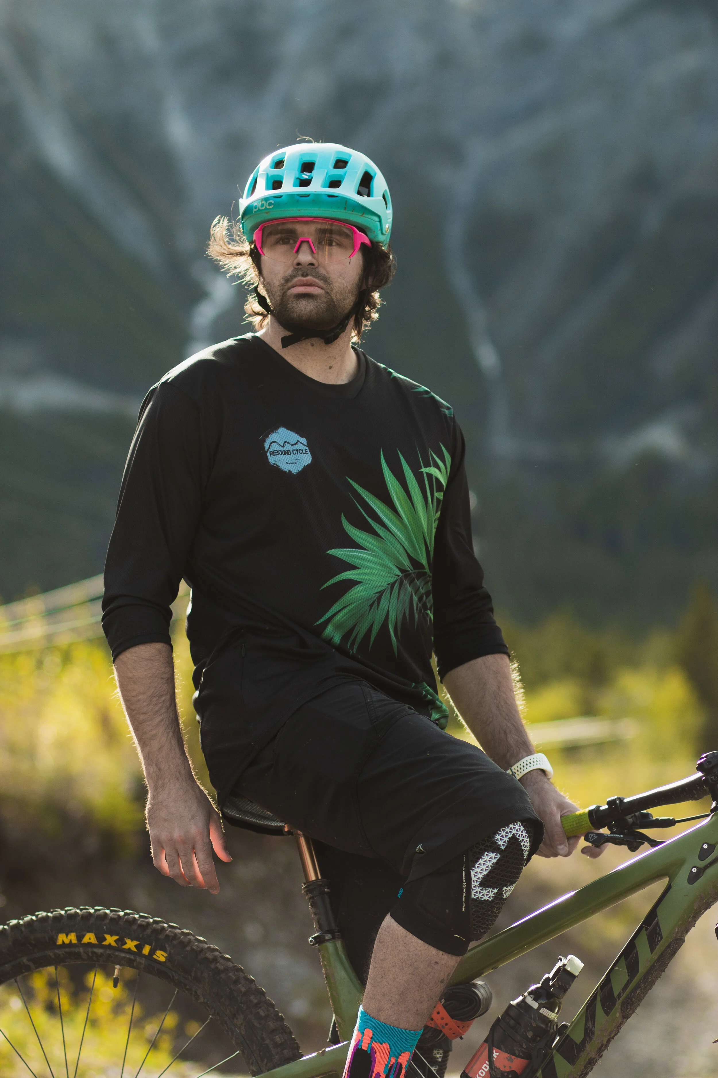 A man with long hair and a beard wearing a teal bike helmet, pink sunglasses, a black shirt with a green leaf design, and colorful socks, sitting on a mountain bike outdoors with blurred trees and sunlight in the background.