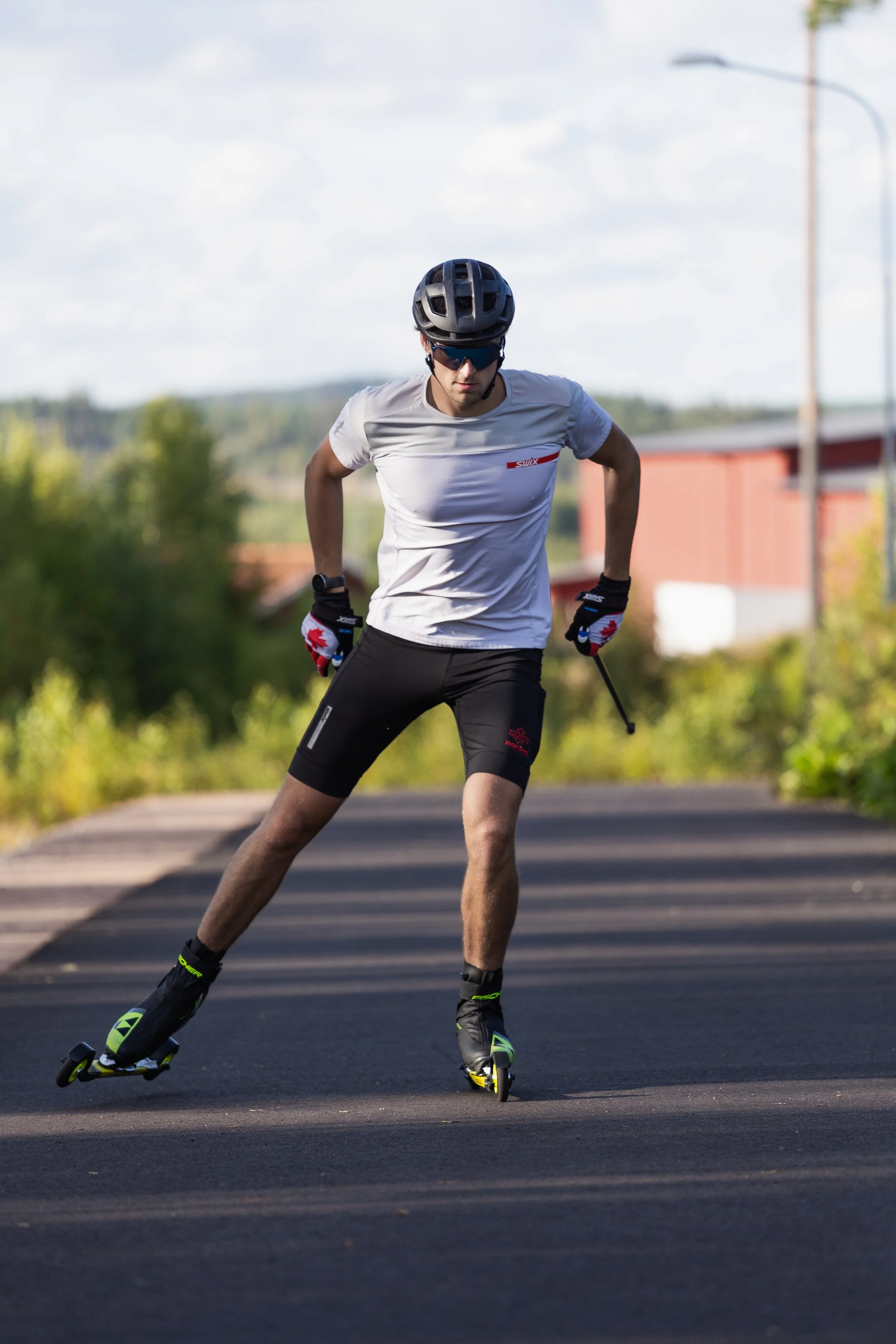 A man wearing a helmet and sunglasses rollerblading on a paved road outdoors with greenery and a building in the background.