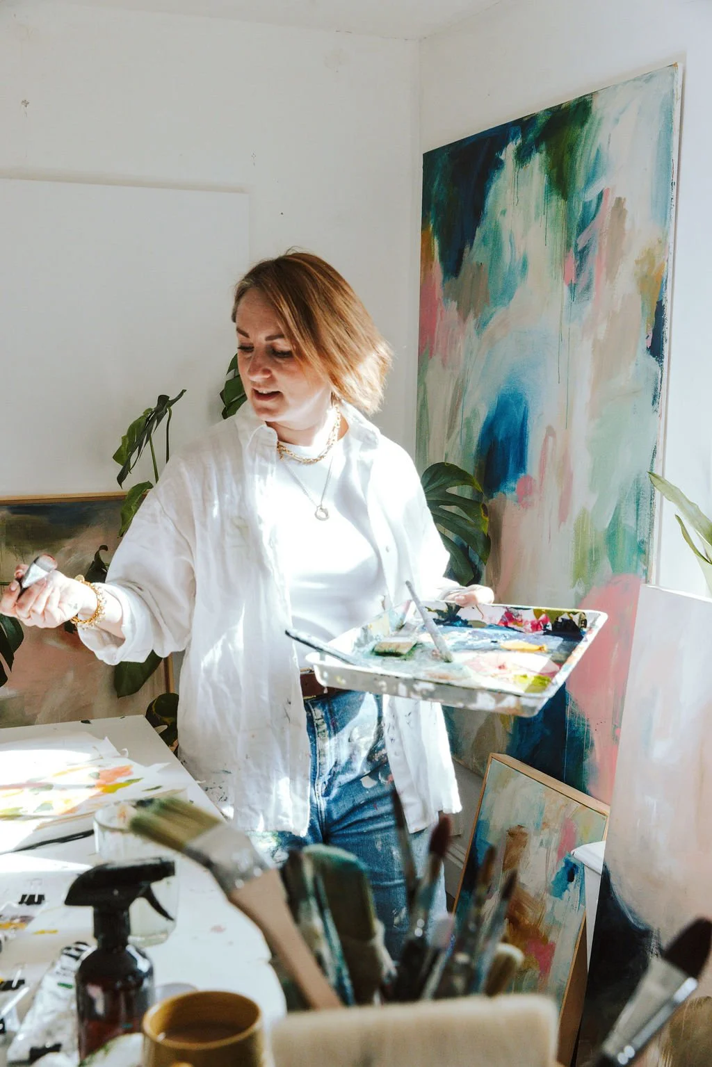 An artist woman in a white shirt and jeans holding a palette with paints and brushes, standing in her studio with abstract paintings and plants around her.