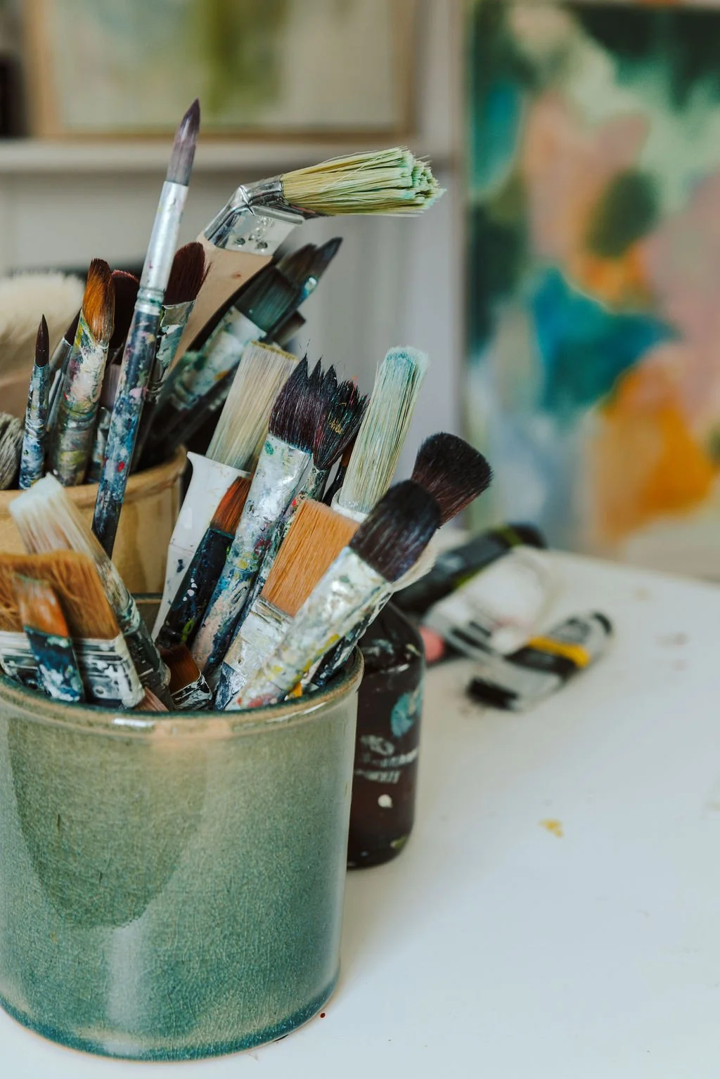 A container holding various used paintbrushes with paint on them, placed on a white table next to paint bottles and a blurred colorful painting in the background.