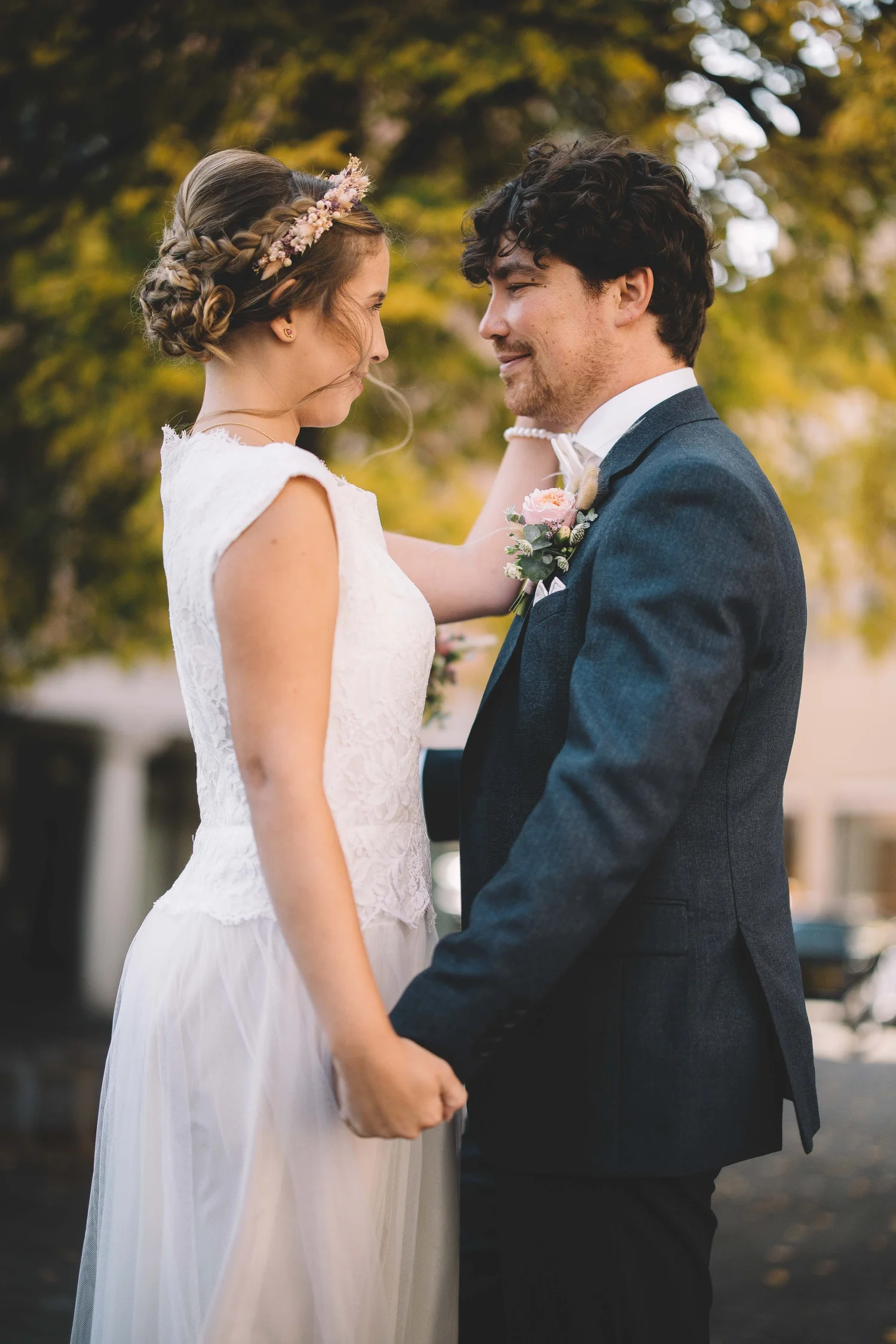 Ein Brautpaar bei einer Hochzeit, stehend vor Bäumen, die Blätter in Herbstfarben haben. Die Braut trägt ein weißes Spitzenkleid mit Zopf-Haarfrisur und Blumenkranz, der Bräutigam trägt einen dunklen Anzug mit Krawatte und Anstecker.