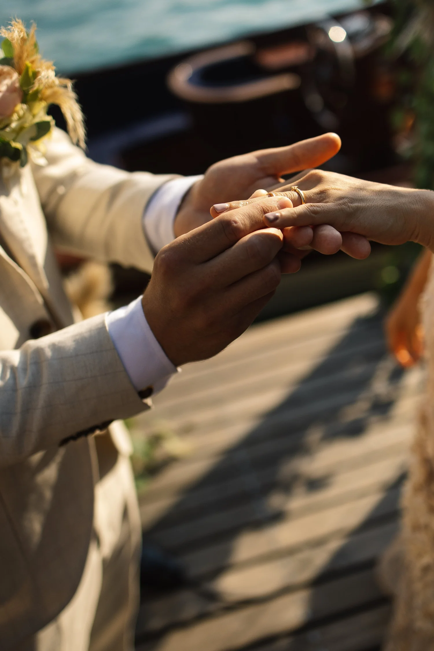 Ein Mann in einem cremefarbenen Anzug hält die rechte Hand einer Frau, die einen Ehering trägt, bei einer Zeremonie am Wasser.