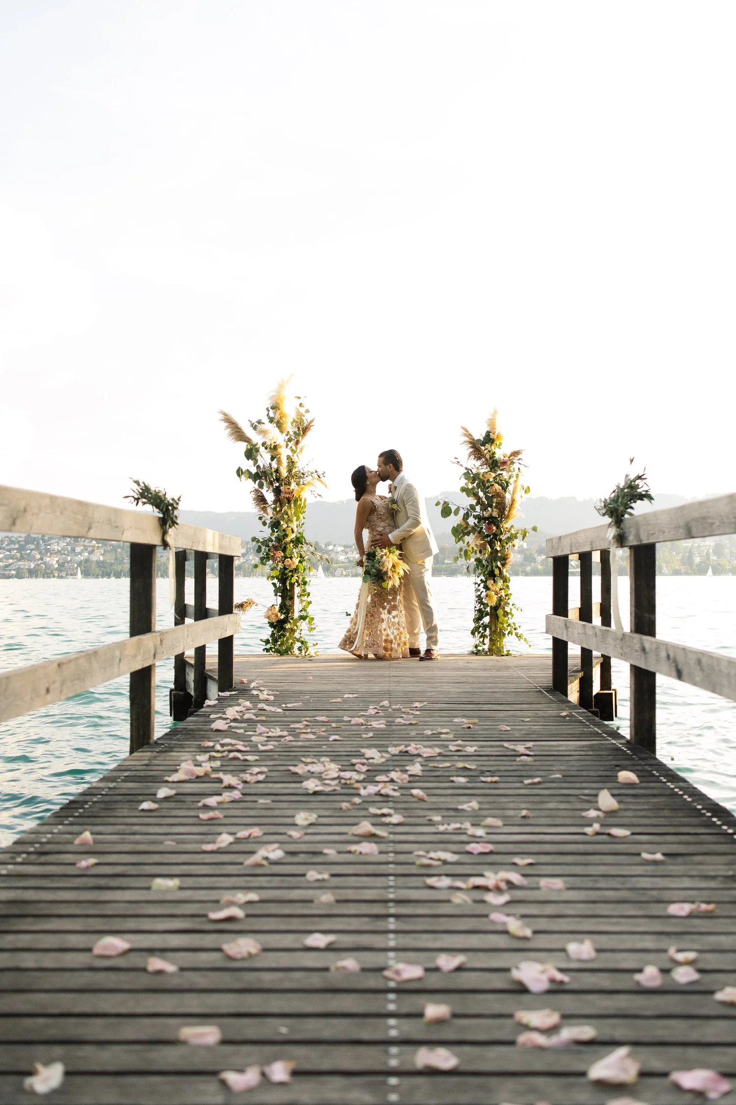 Ein Hochzeitsbild eines Paares, das sich auf einem Steg am Wasser küsst, mit Blumenarrangements im Hintergrund und Rosenblüten auf dem Boden, bei Sonnenuntergang im Freien.