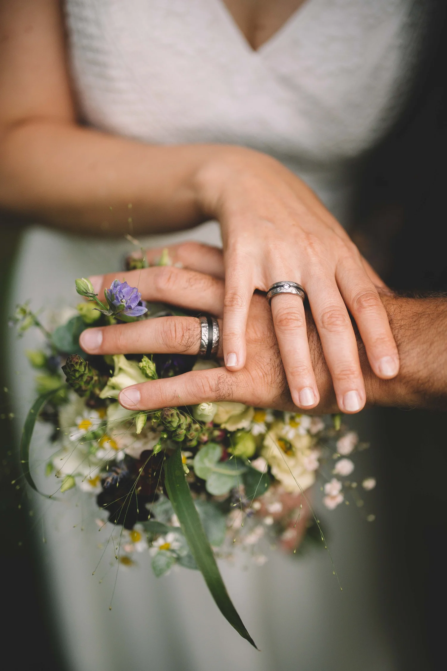 Nahaufnahme von zwei Händen mit Eheringen, die übereinander auf einem Blumenstrauß liegen.