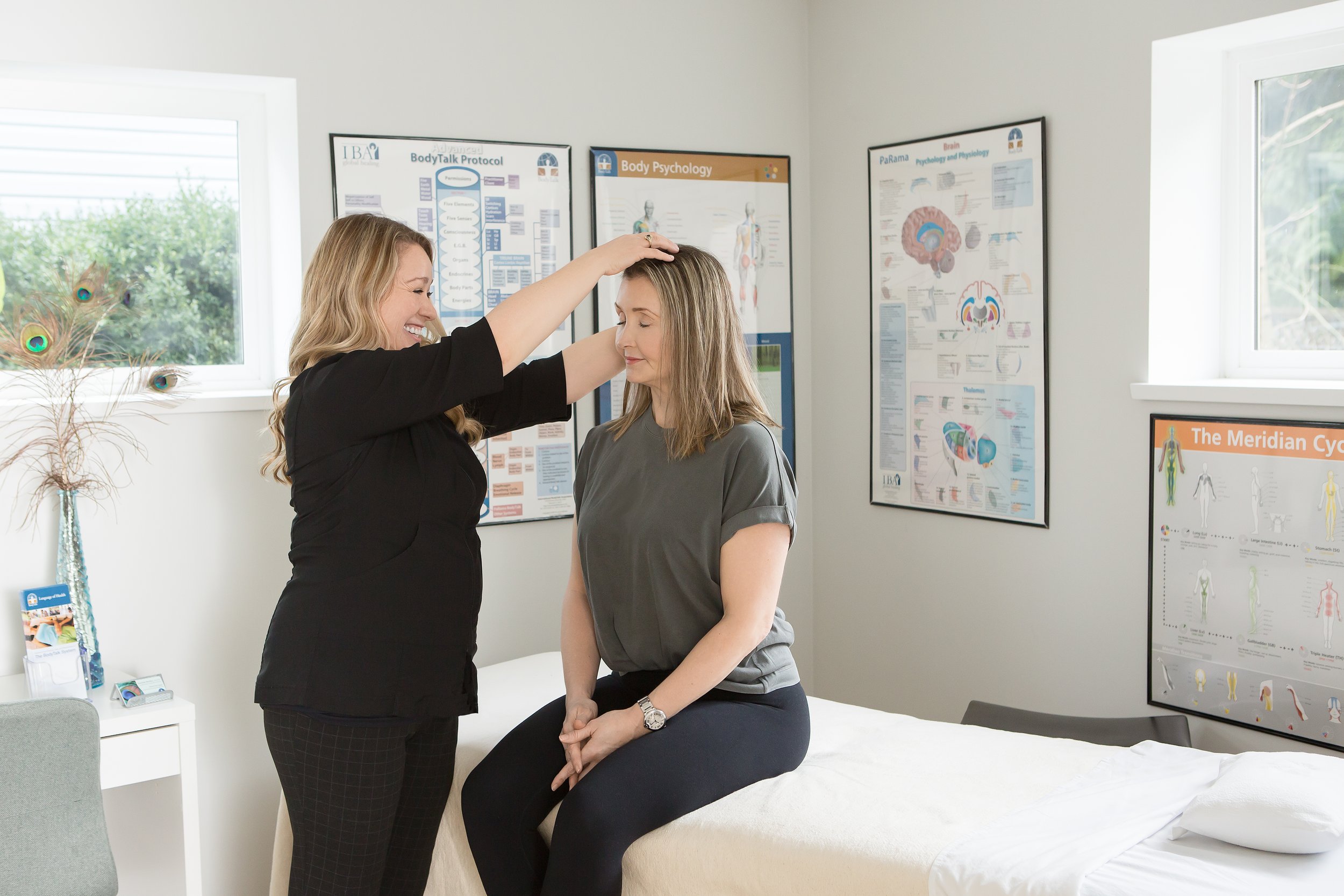 A woman in a gray shirt sitting on a medical examination table with her eyes closed, while a healthcare professional stands beside her, gently placing her hand on the woman's head in a supportive gesture. The room has educational posters on the wall 