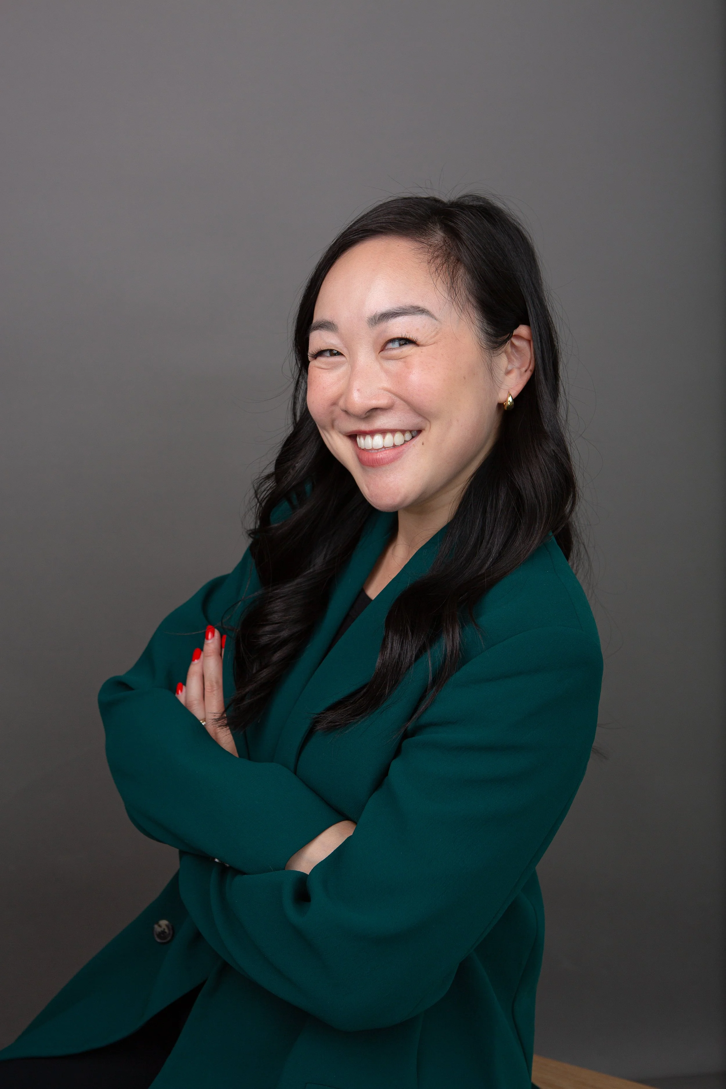 A woman in a green blazer smiling, with arms crossed, against a gray background.