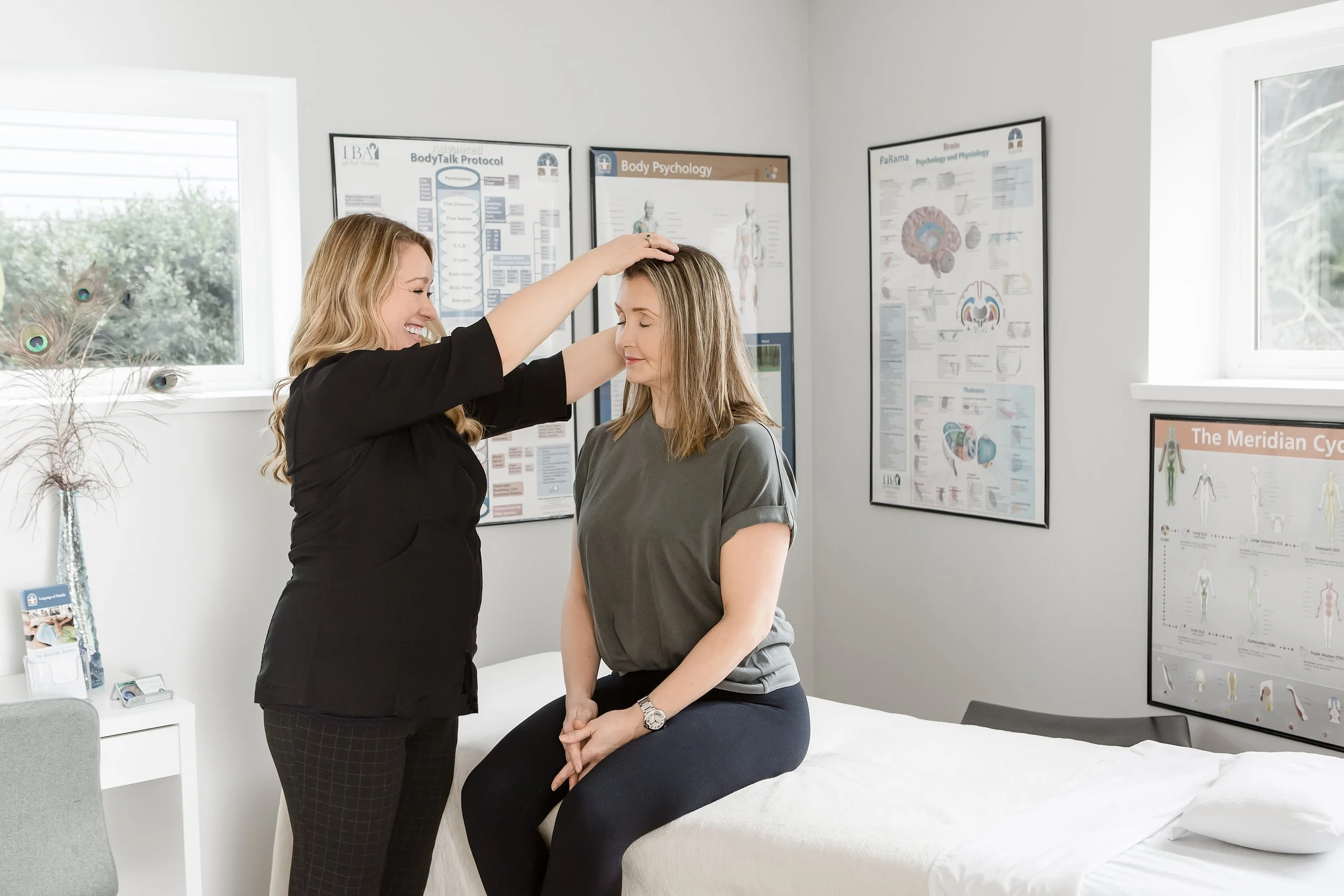 A woman is seated on an examination table in a medical office, with her eyes closed and hands clasped on her lap. Another woman, possibly a healthcare professional, is gently placing her hand on the patient's head as part of a calming or therapeutic 