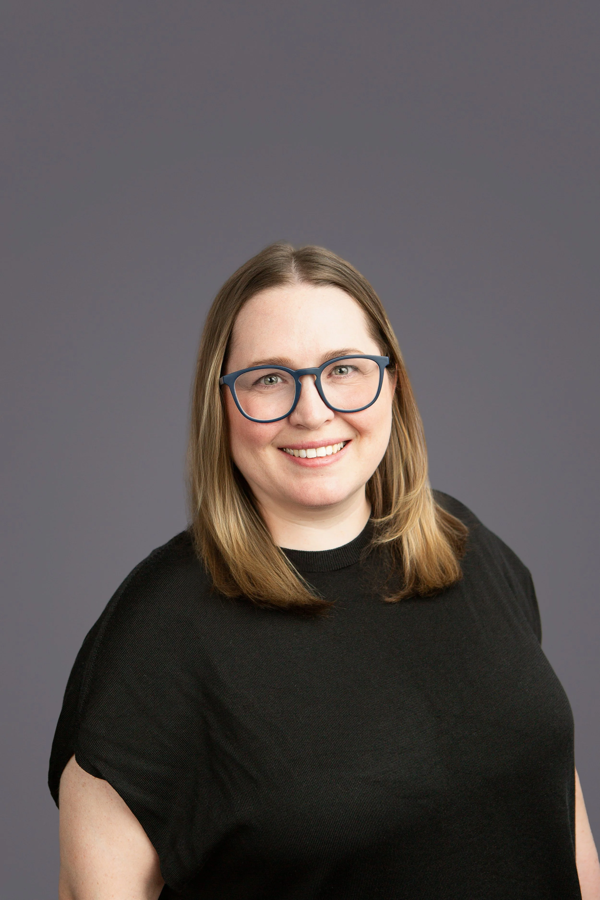 A woman with shoulder-length light brown hair, wearing glasses with thick frames, smiling, dressed in a black shirt, against a plain gray background.