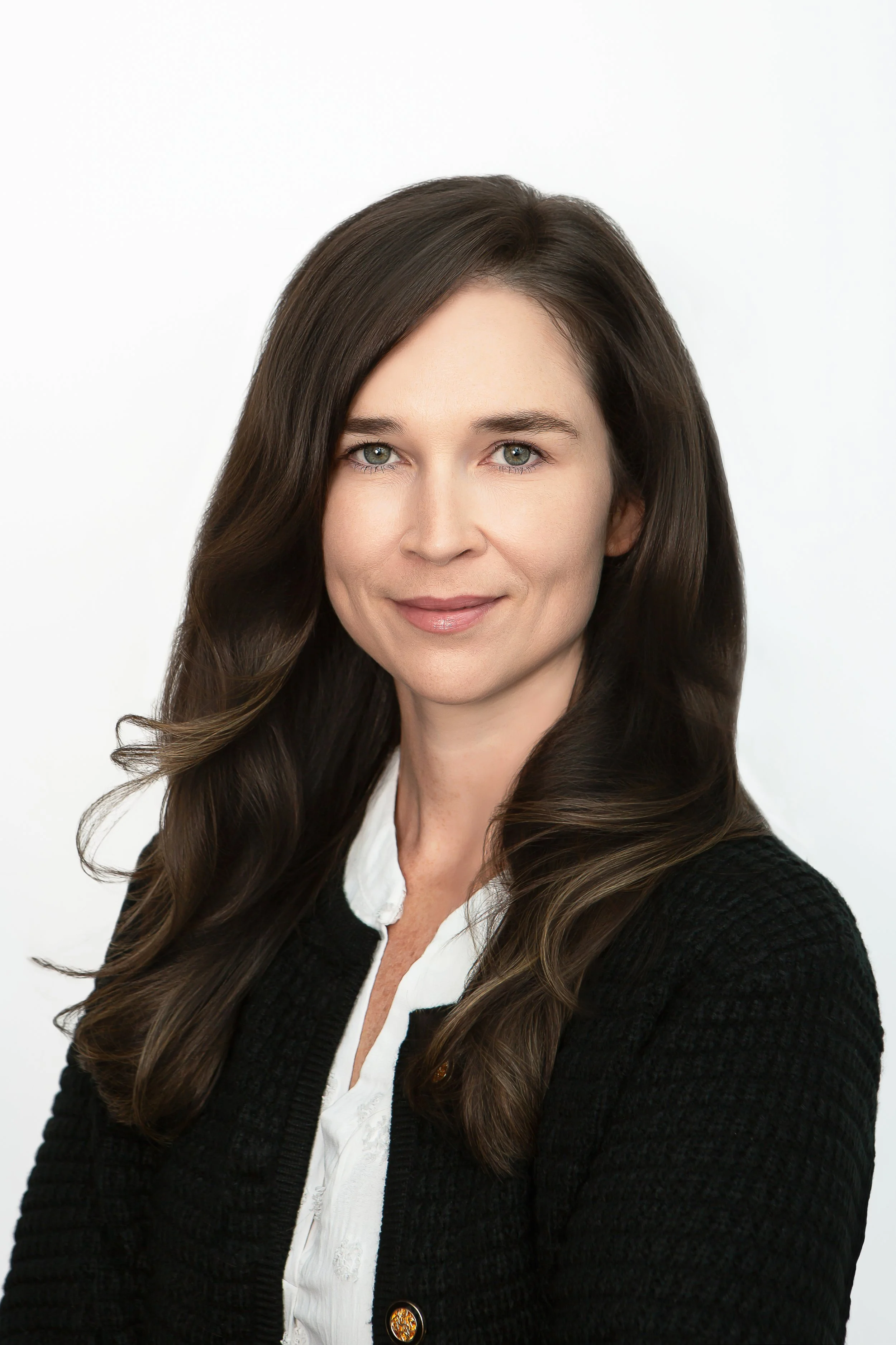 A woman with long, wavy brown hair wearing a black cardigan and white blouse, smiling, against a plain white background.