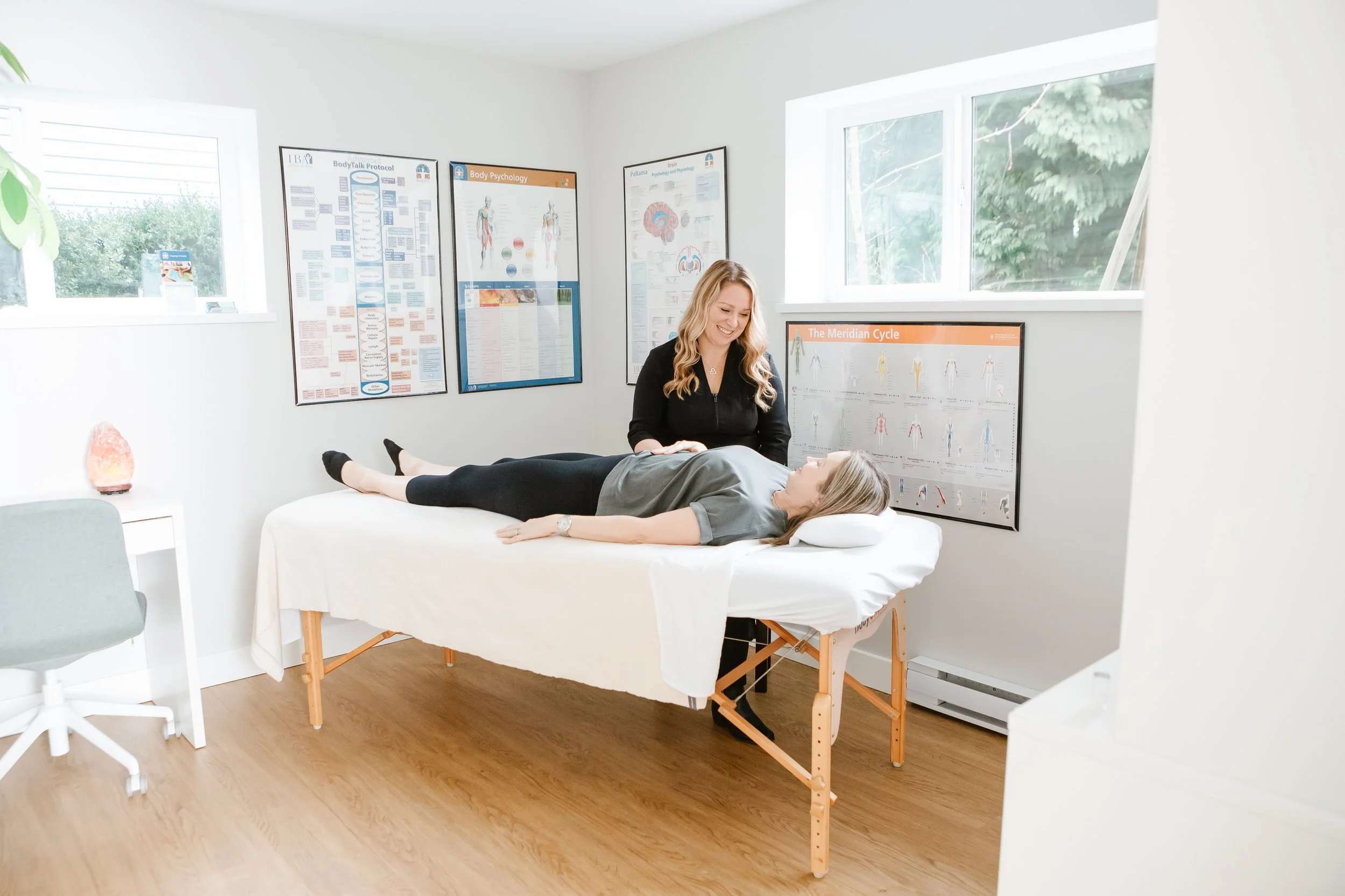 A therapy session with a woman lying on an examination table and a therapist smiling at her. The room has medical posters on the wall and bright natural lighting from the window.