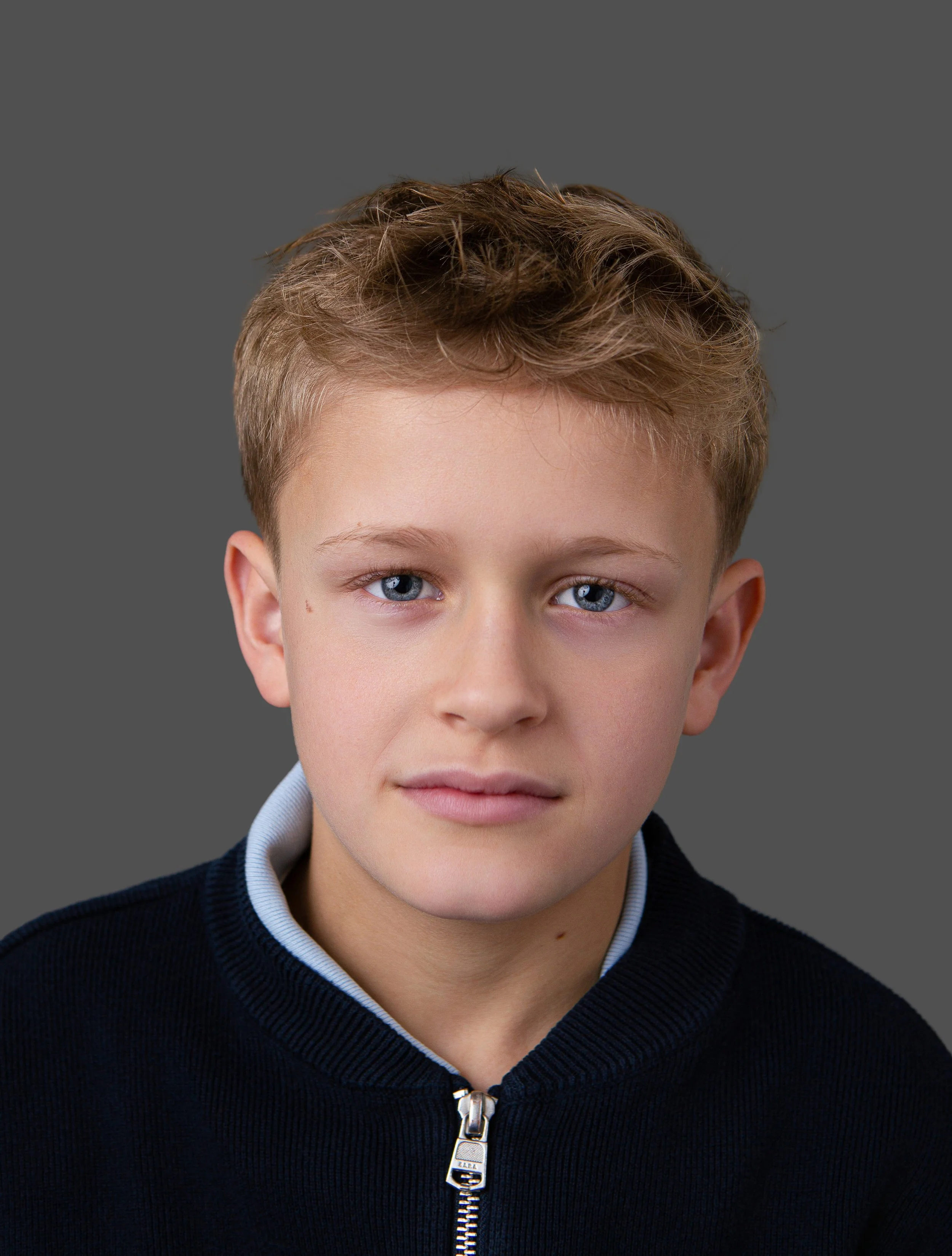 Close-up of a young boy with blue eyes and light brown hair, wearing a black zip-up sweater with a light blue collar, against a gray background.