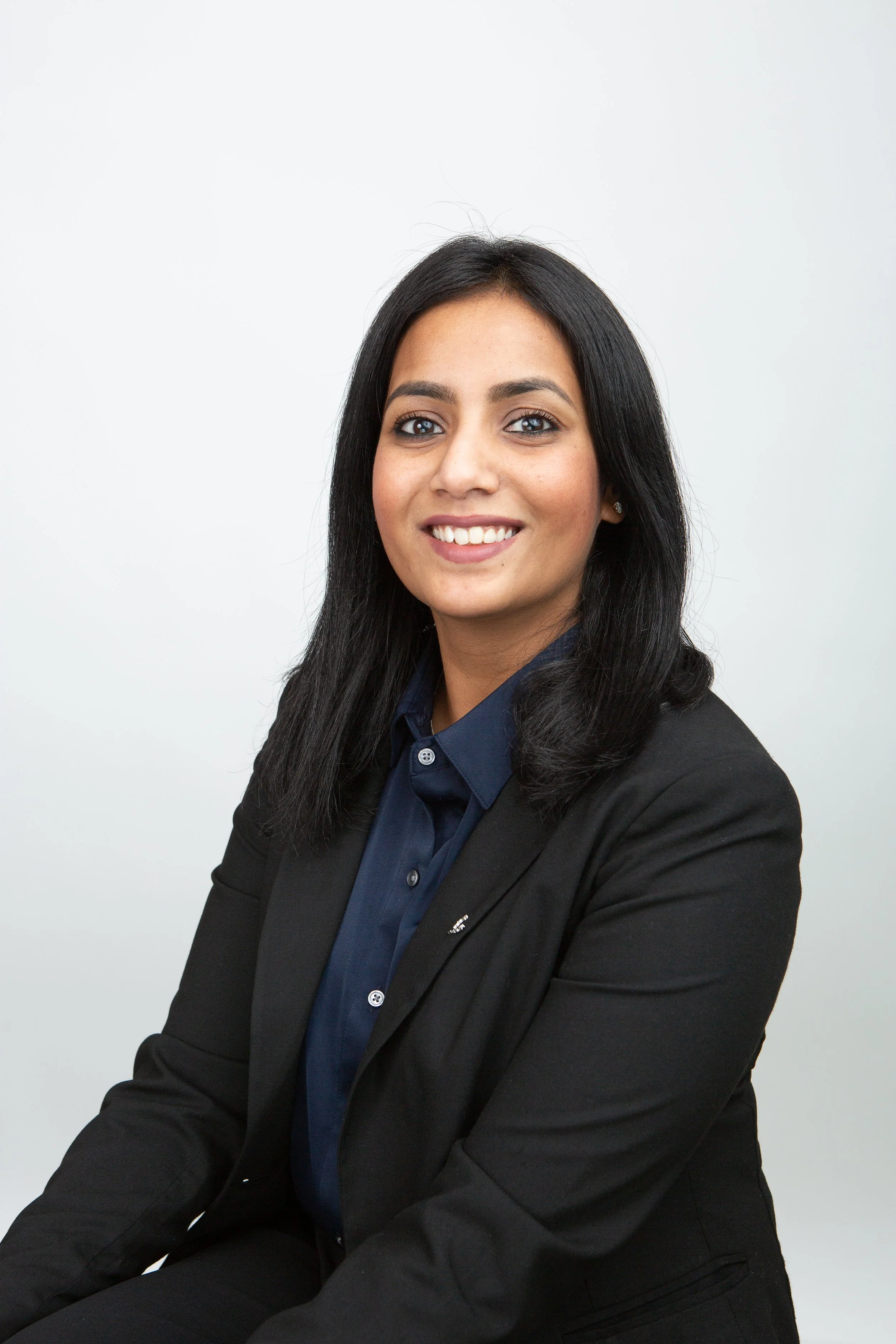 A professional woman with long dark hair, wearing a navy blue shirt and black blazer, smiling and seated against a plain white background.