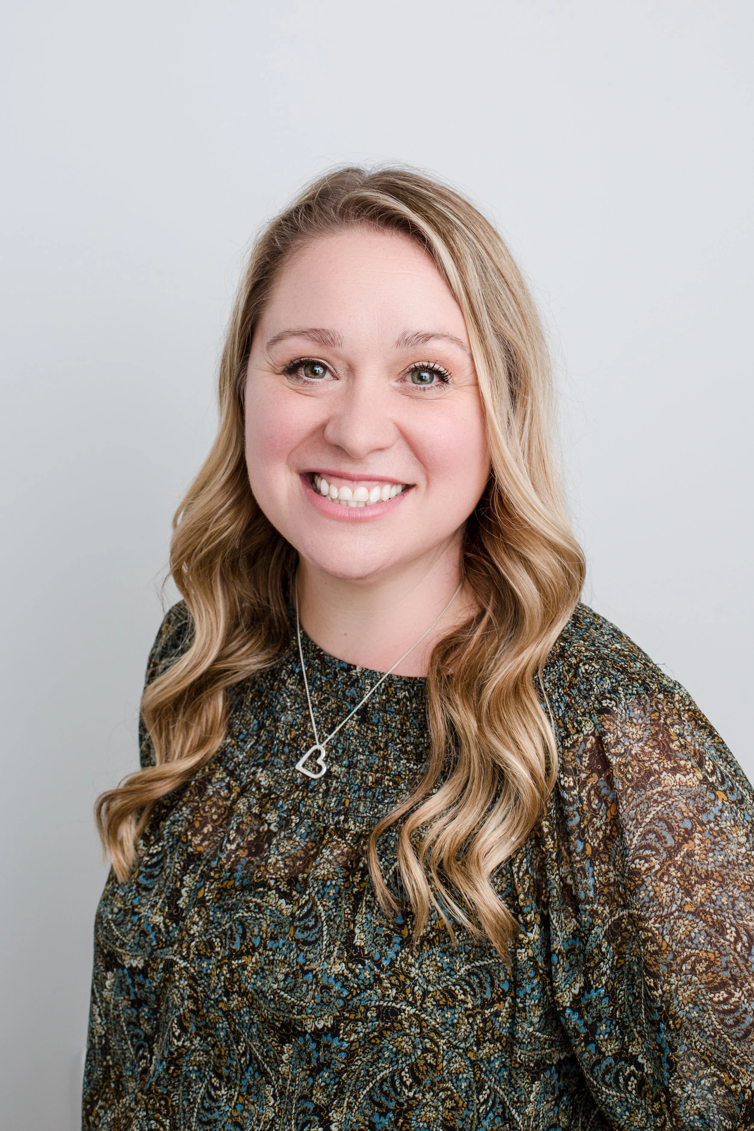 A woman with long wavy blonde hair, smiling, wearing a patterned dress and a silver necklace with a heart pendant, against a plain white background.