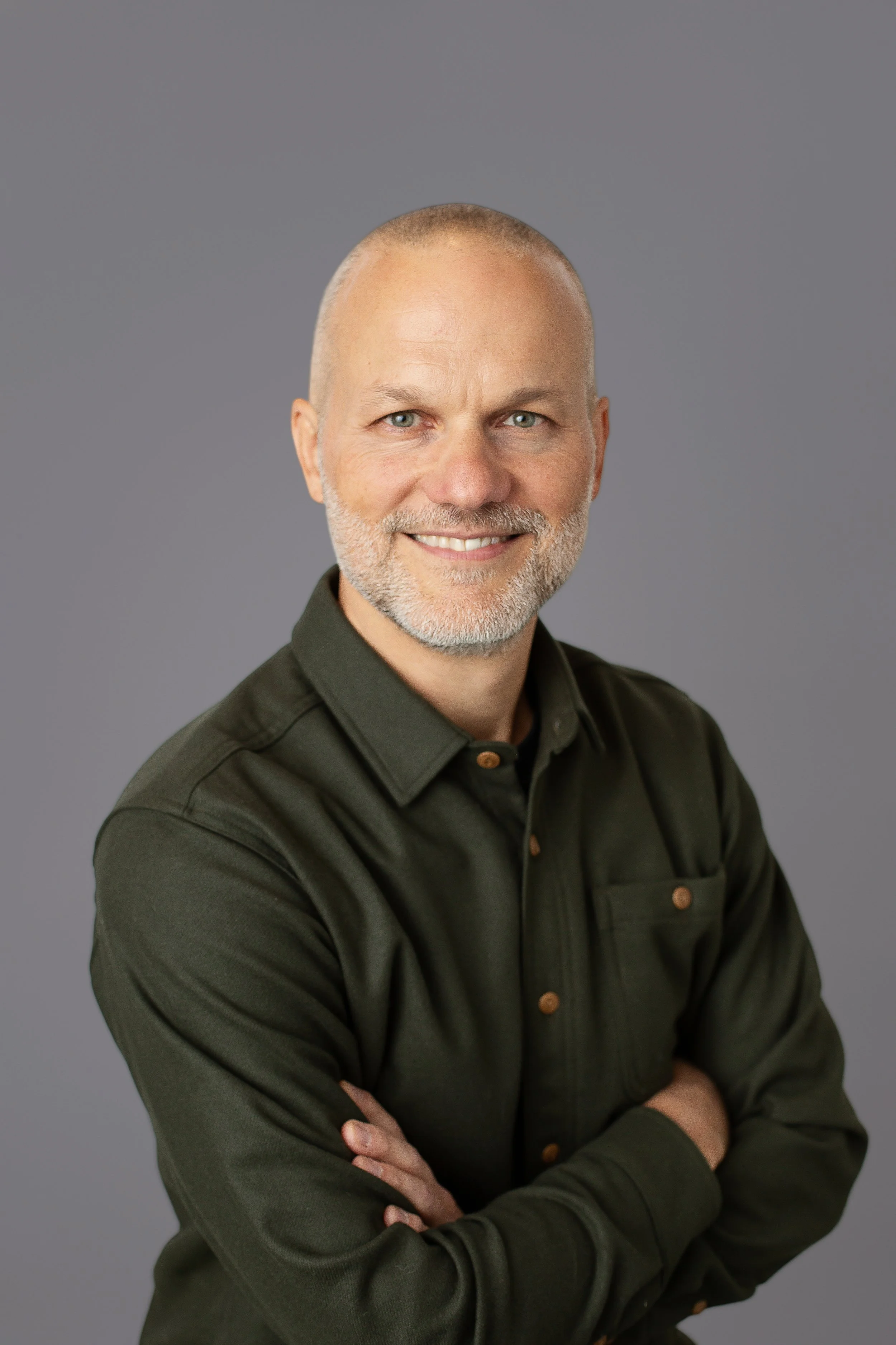 A middle-aged man with a beard and short hair, wearing a dark green shirt, smiling with arms crossed in front of a plain grey background.