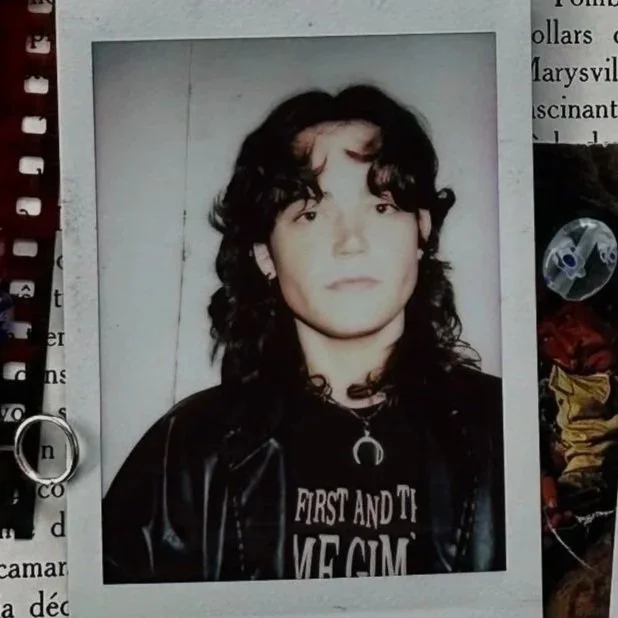 A young person with dark, curly hair and light skin, wearing a black leather jacket and a necklace with a crescent moon charm, standing against a plain background.