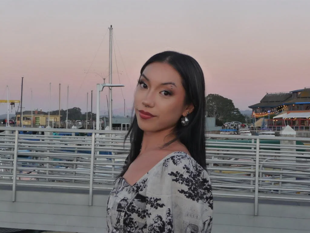 A woman with black hair and earrings standing near a marina with boats and buildings in the background during dusk.