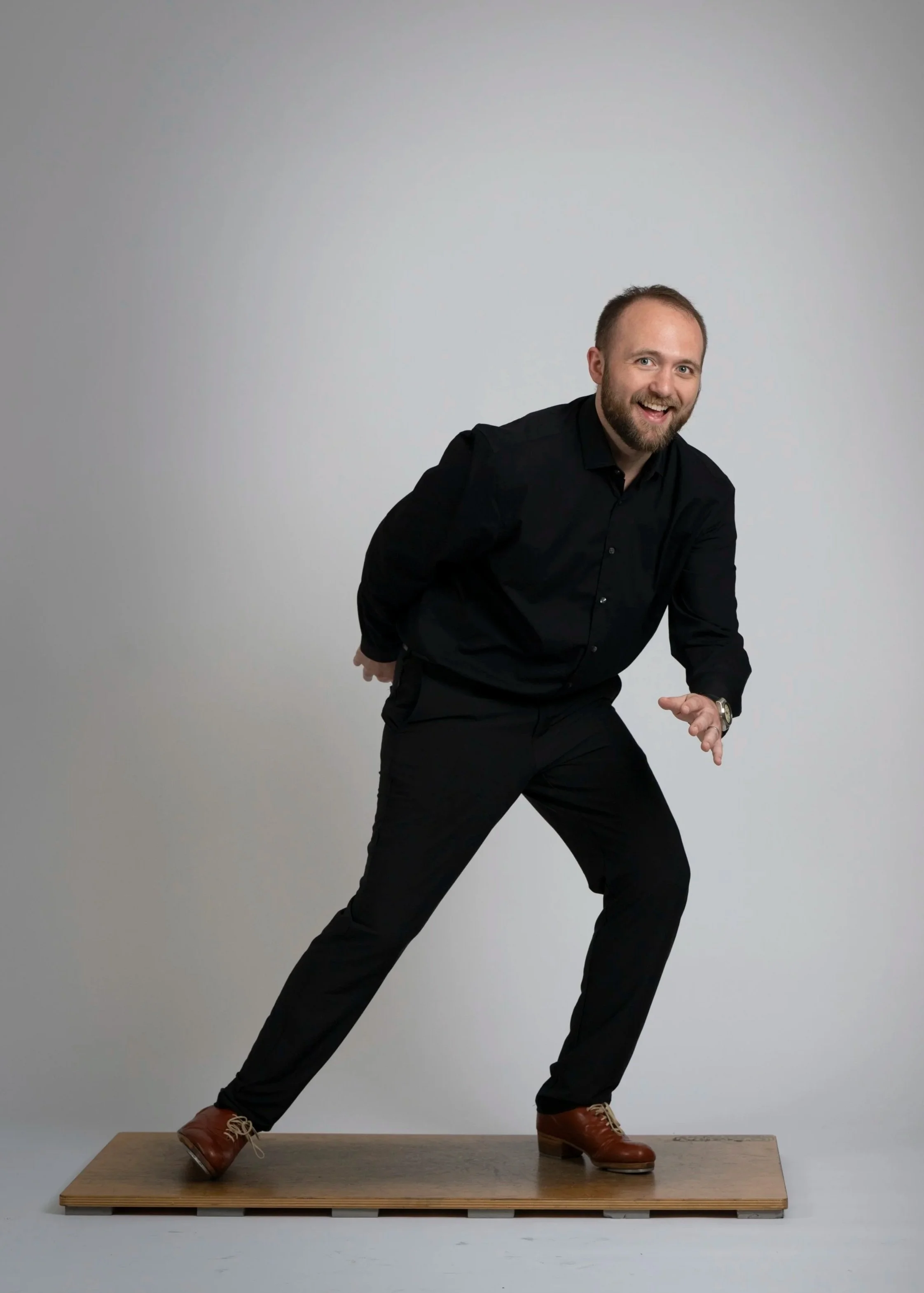 A man with a beard and short hair, dressed in a black shirt and black pants, is standing on a wooden platform against a plain gray background. He is bent forward with one hand on his knee and the other hand extended, smiling and looking at the camera