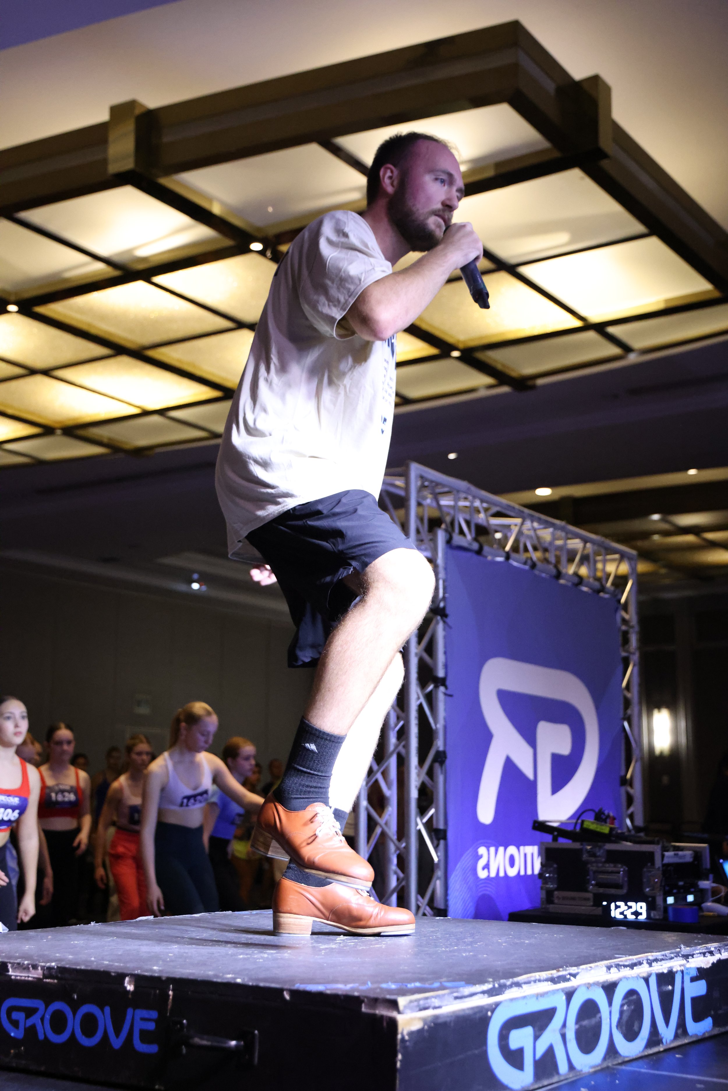 A man with a beard holding a microphone on a stage, wearing a white shirt, black shorts, and brown boots, with a group of girls standing behind him, in a room with a large ceiling and stage lights.