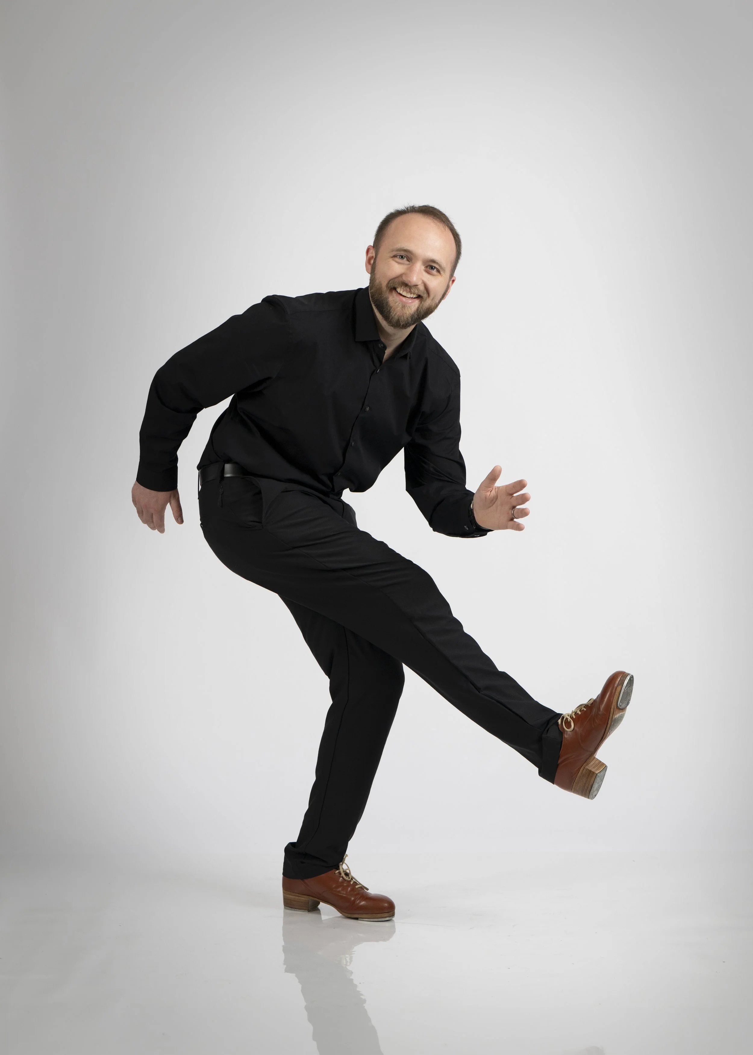 A man with a beard is wearing a black shirt and pants, brown shoes, and is smiling while balancing on one foot with his other leg raised and his hands in a playful pose.
