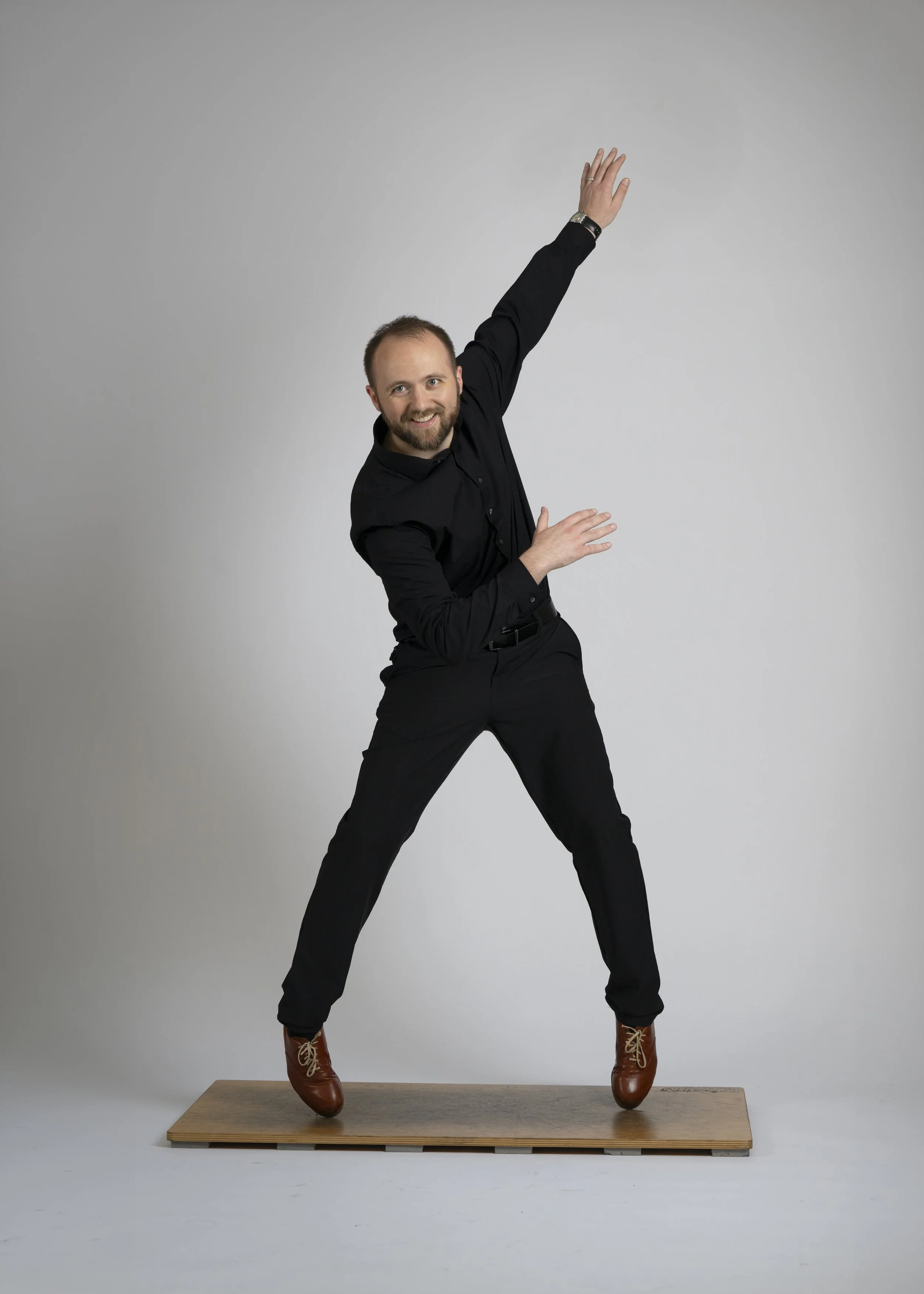 A man with a beard in black clothing and brown boots balancing on a wooden platform in a pose with one arm raised and one arm bent, against a plain gray background.