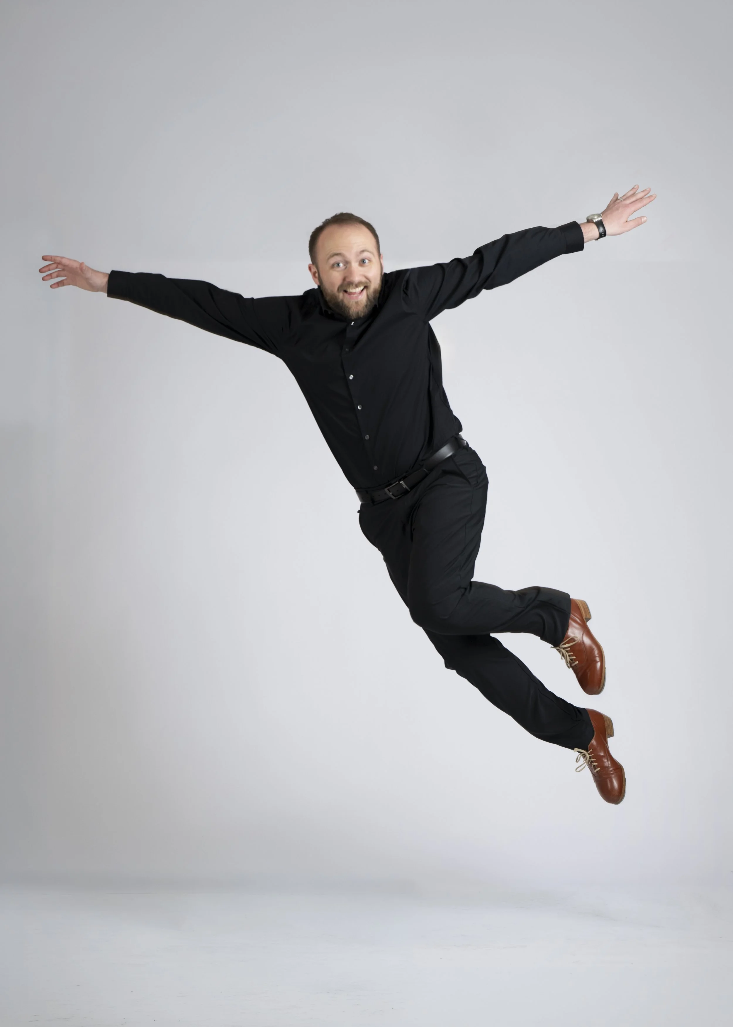 A man in black shirt and pants, brown shoes, smiling and with arms outstretched, appears to be jumping or floating in front of a plain white background.